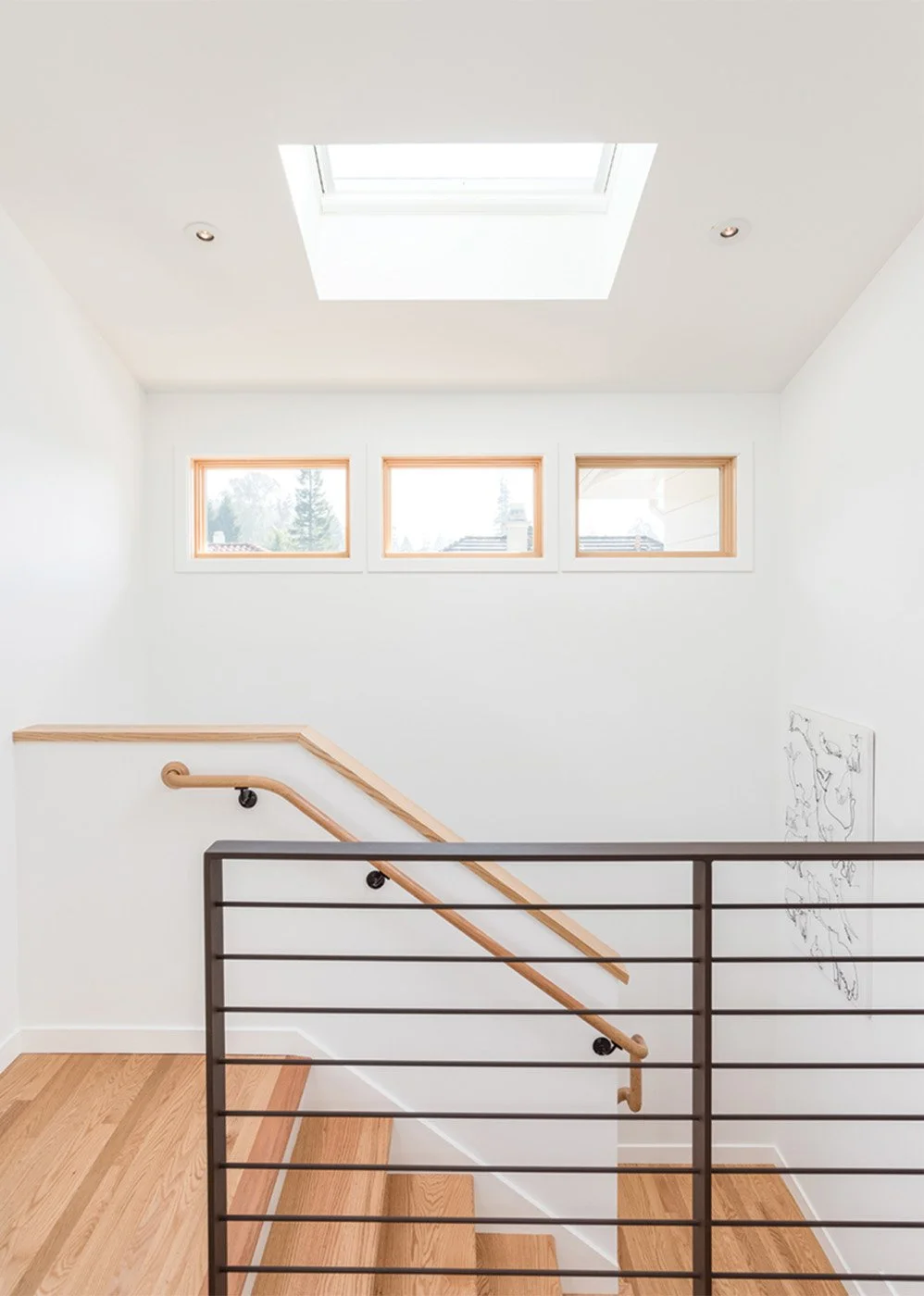 Stairwell with natural wood railing, black horizontal metal balusters, and skylight above illuminating the white walls and oak flooring