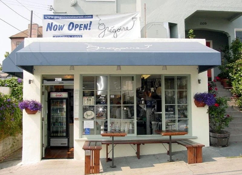 Front exterior of Grégoire Piedmont in a historic Julia Morgan building, with blue awning, open kitchen visible through the windows, and outdoor bench seating along the sidewalk.