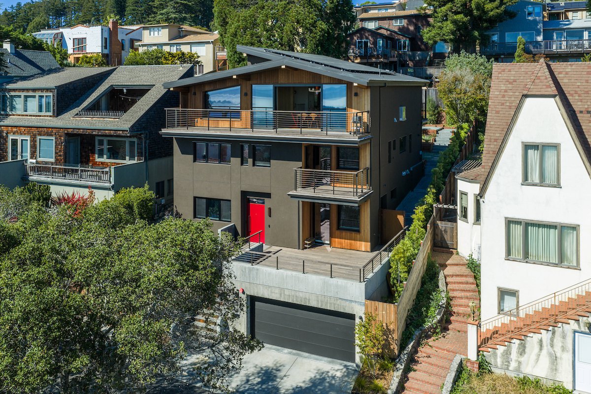 The stepped hillside structure is organized around multiple terraces and balconies, extending interior living spaces outward. Dark stucco, natural wood siding, and metal railings create a warm, modern material contrast.