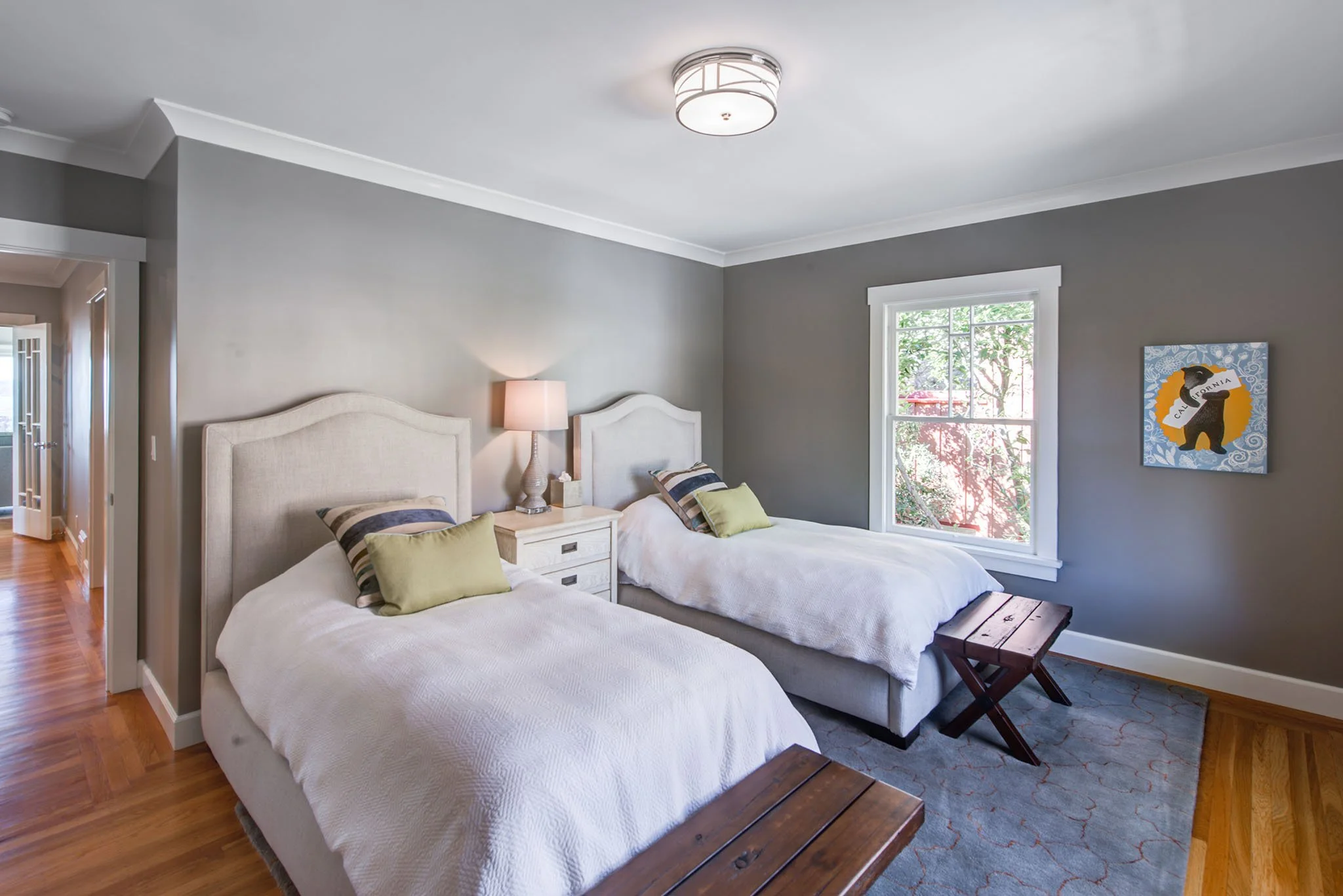 Updated guest bedroom with two upholstered twin beds, gray walls, and warm wood floors. A large window and simple finishes echo the calm palette used throughout this Berkeley remodel.