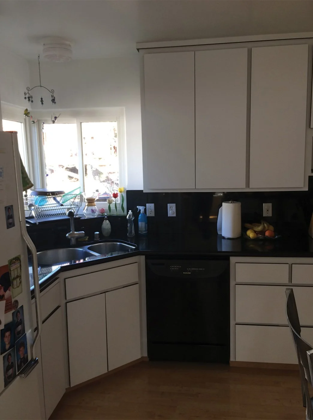 Before photo of the original kitchen with white cabinets, dark counters, older sink under the window, and black dishwasher before Levitch updated finishes and fixtures.