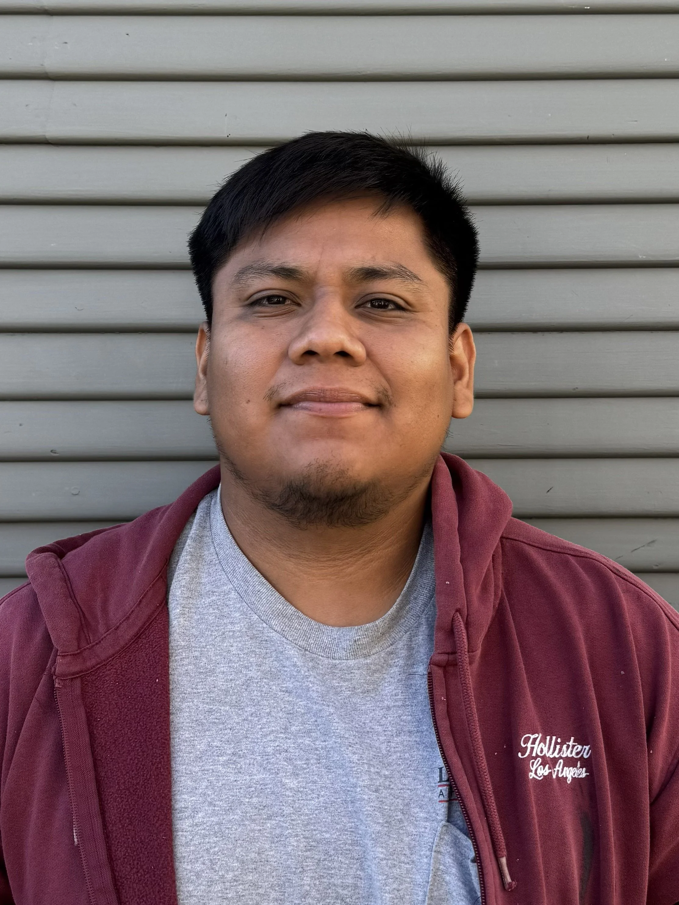 A young man with short black hair and light brown skin, wearing a gray T-shirt and maroon hoodie, standing in front of a gray metal shutter.