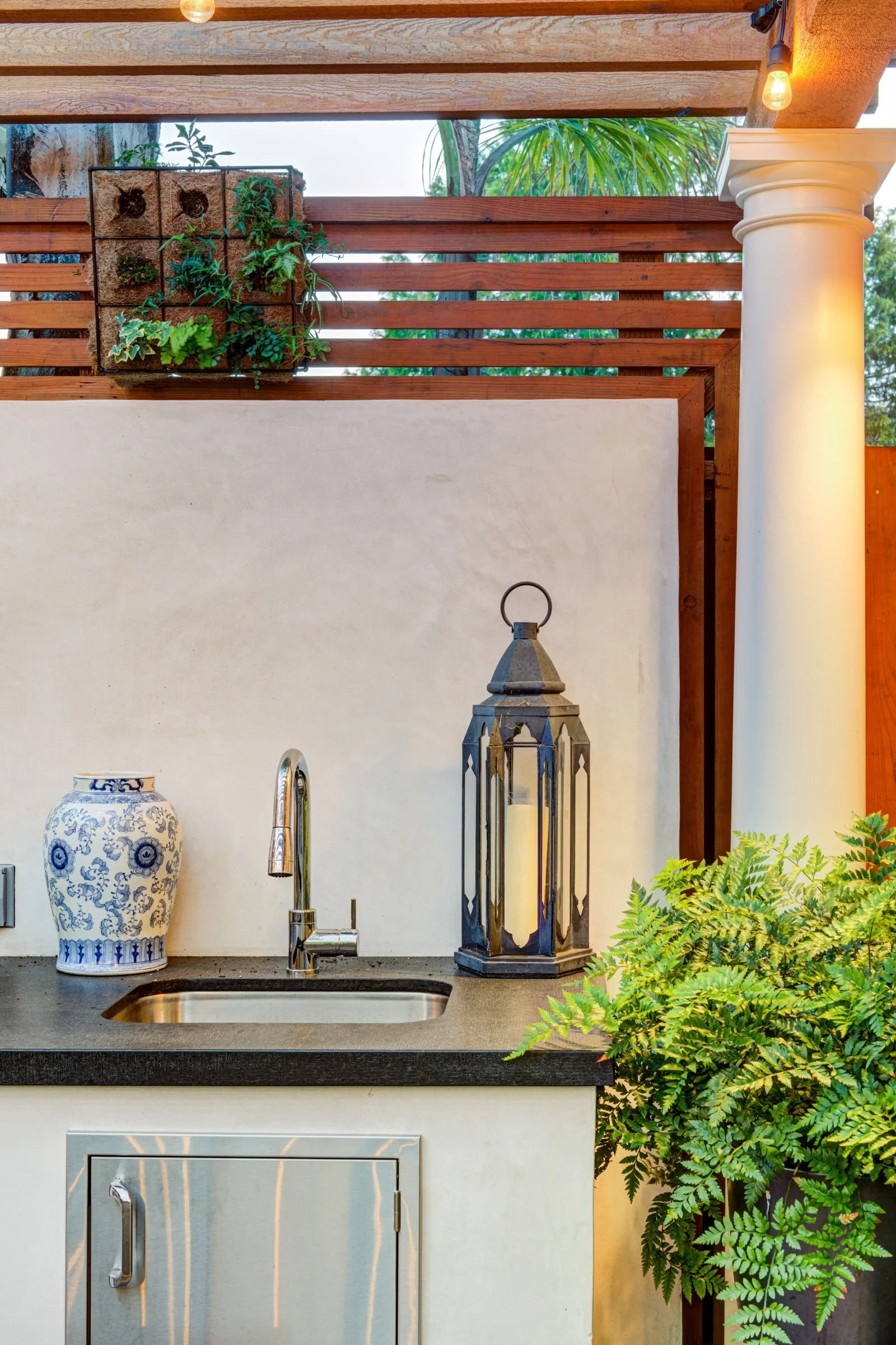 Detail view of the outdoor kitchen work zone highlighting a stainless steel sink, lantern-style lighting, and decorative ceramic accents. Natural materials and greenery soften the space while supporting food prep and outdoor use.