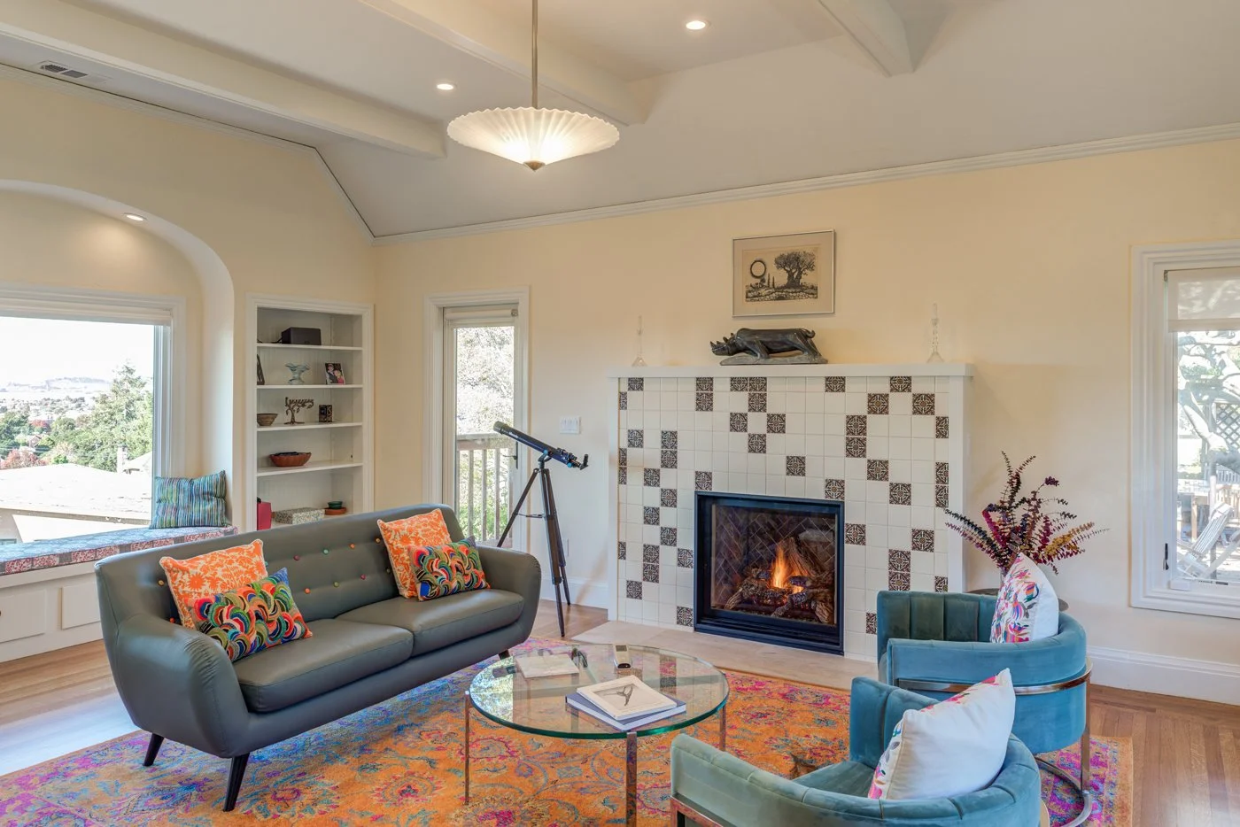 Bright living room after remodel with a new gas fireplace and patterned tile surround, built in shelving, large windows, and modern seating on a colorful rug.