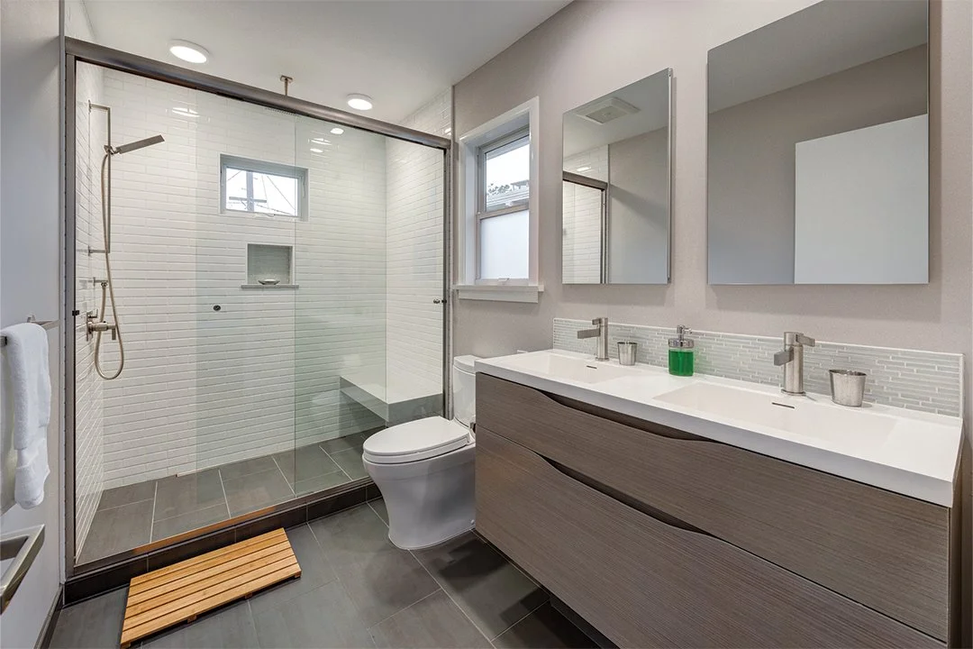 Primary bathroom with floating double vanity, two mirrors, gray tile floor, and a glass shower with bench and built-in niche, completed as part of the new suite addition.