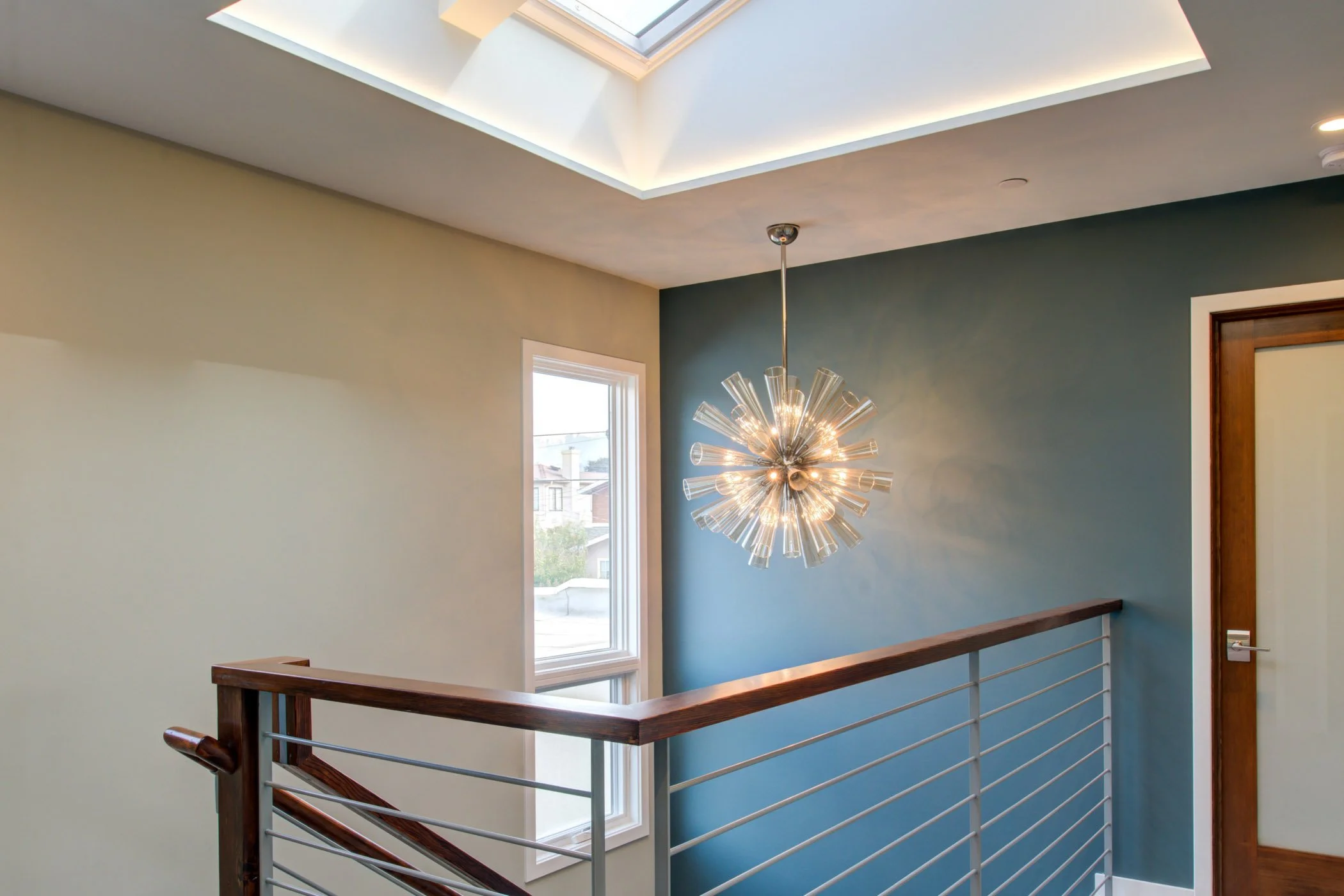 Detail of the bright stair landing with a starburst chandelier, teal feature wall, tall window, and slim cable railing that keeps the new second-story circulation light and open.