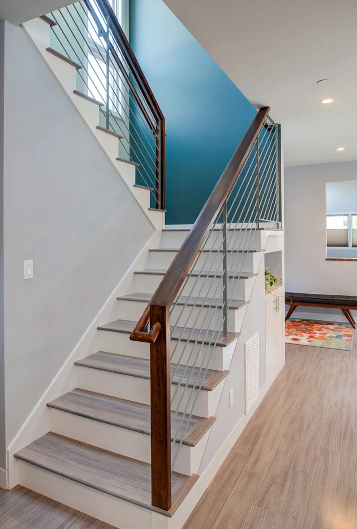 Minimal footprint stair to the new second story with wood handrail and cable guardrail, grey treads, built-in storage below, and a bold blue accent wall for a warm modern feel.