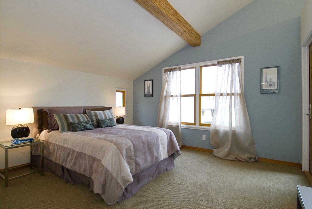 Primary bedroom with vaulted ceiling, exposed wood beam, soft blue accent wall, and large windows that maximize daylight while creating a calm and comfortable private space.