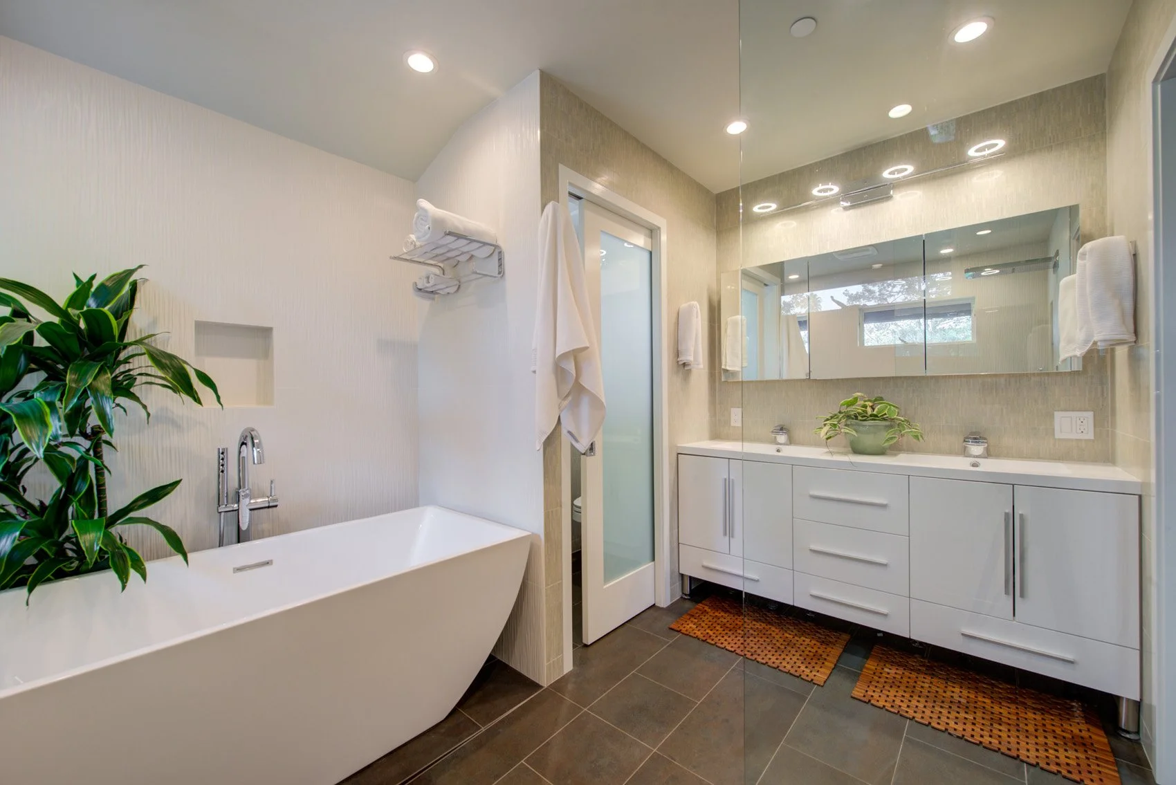 Primary bathroom with a freestanding soaking tub, long floating vanity and full-width mirror, glass shower enclosure, and warm neutral finishes that make the space feel calm and bright.