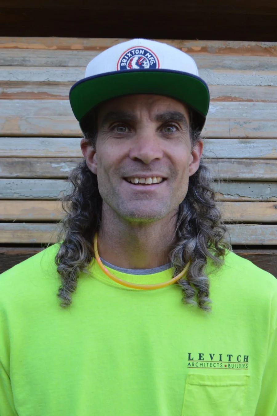 Man with long curly gray hair wearing a white and blue Brixton MEC cap, bright yellow shirt with 'LEVI TCH ARCHITECTS BUILDERS' on it, standing in front of a wooden wall.