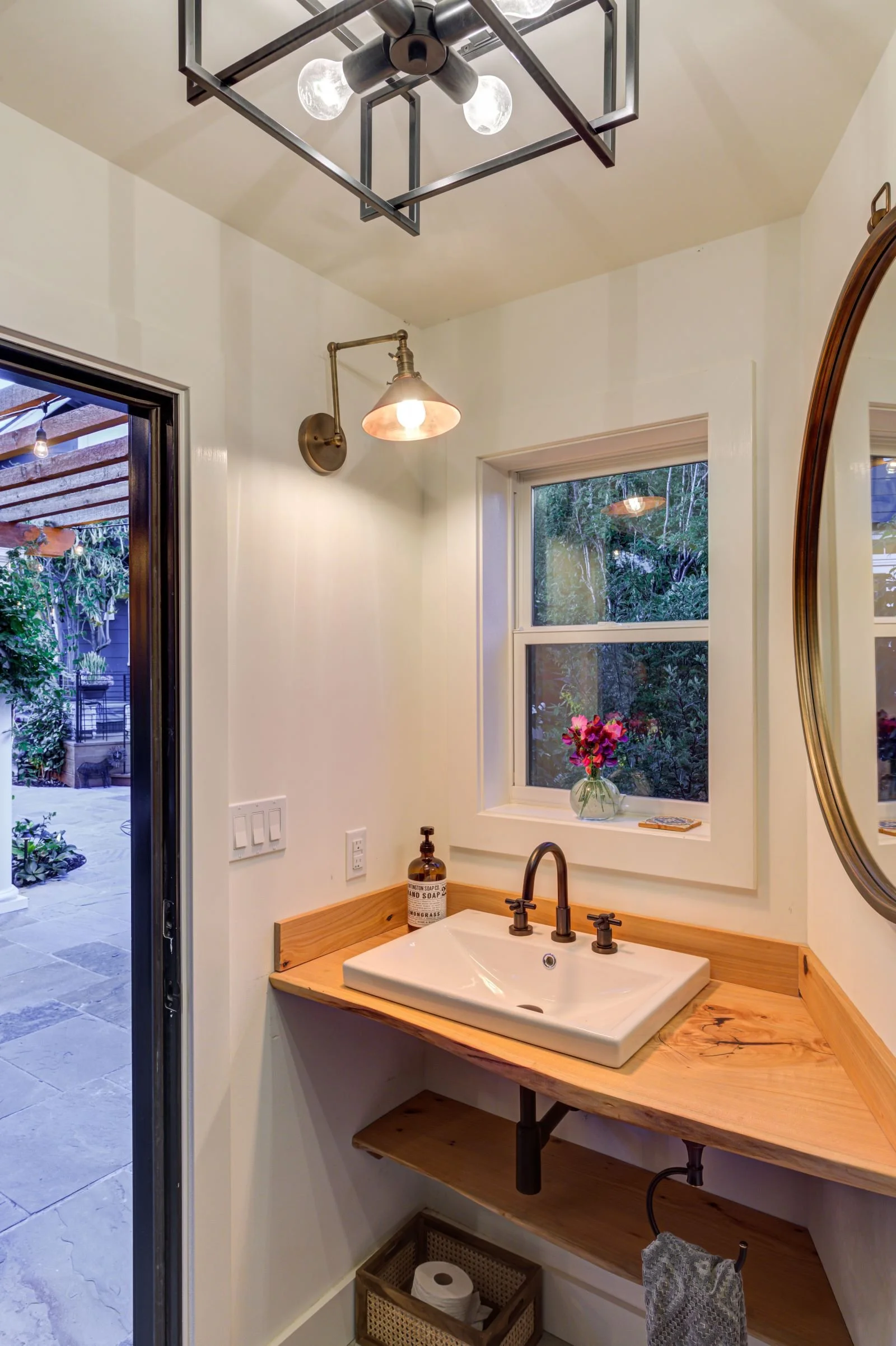 Compact powder room featuring a custom live-edge wood vanity with an integrated backsplash and matte black fixtures. Warm brass and black lighting, paired with a small window, bring natural light and warmth to a functional guest Bathroom.