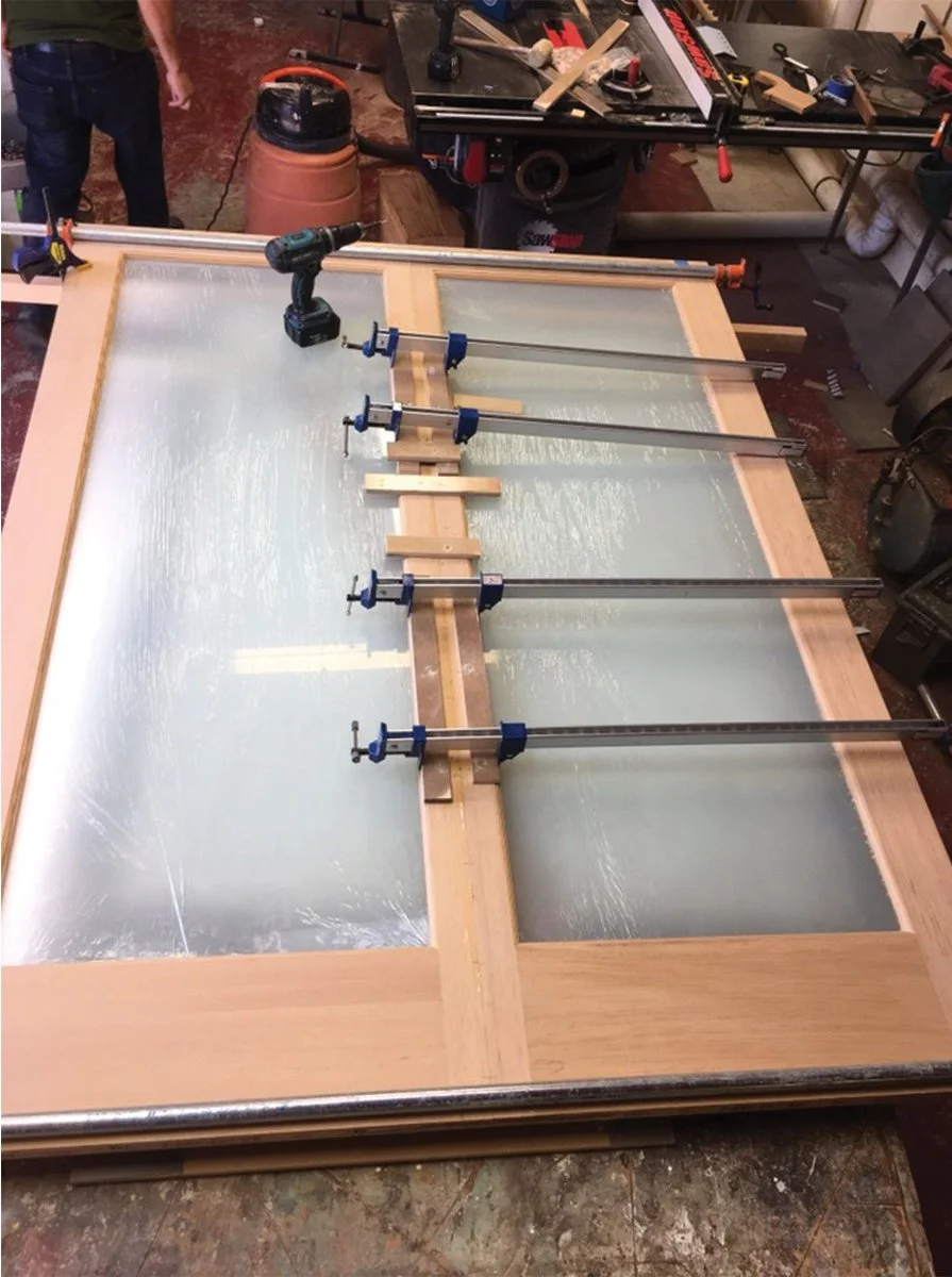 Workshop fabrication of a custom wood framed frosted glass door, clamped on a worktable with tools, being assembled for the new bedroom suite addition built by Levitch.