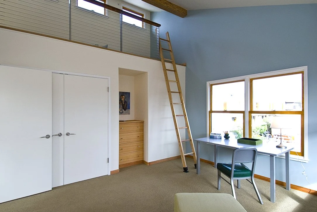 Bedroom with ladder access to a lofted sleeping area, featuring high ceilings, exposed wood beam, and clerestory windows that maximize light in a compact footprint.