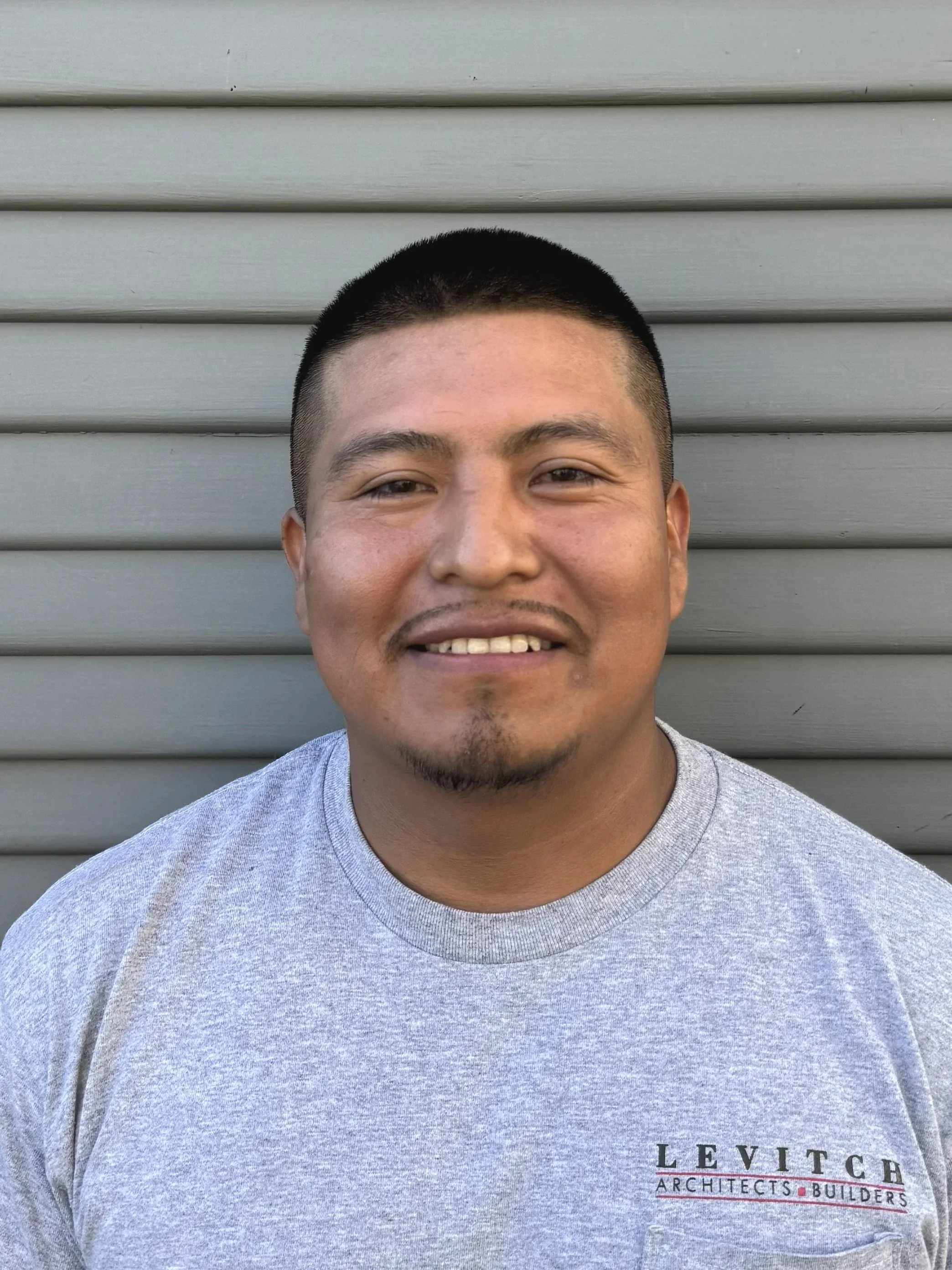 A young man with short black hair and a goatee, wearing a gray T-shirt, smiling in front of a gray horizontal siding wall.