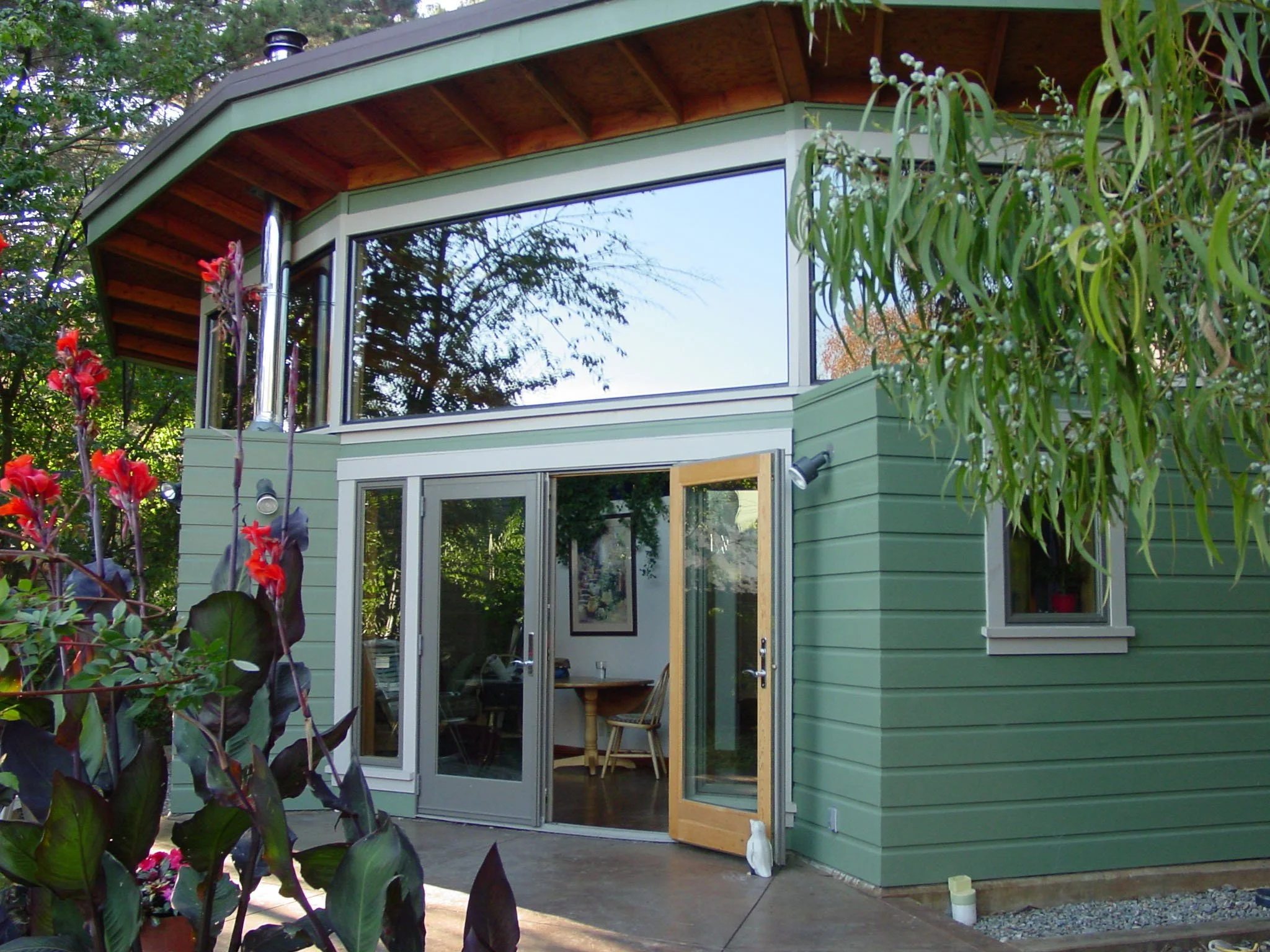 Backyard cottage exterior in the rear garden with green siding, wide glass doors, and high windows under a deep roof overhang, designed by Levitch for daylight and passive solar gain.