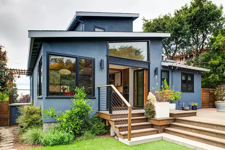 Exterior view of the midcentury home addition in El Cerrito, showing the ground-level ADU and second-story bedroom addition with blue siding, large windows, and a wood deck connected to the backyard.
