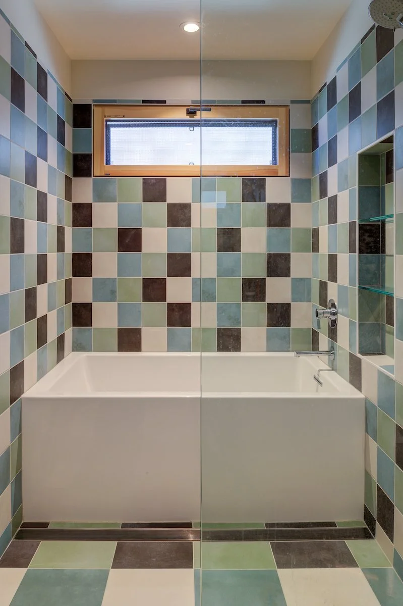 A playful grid of muted green, blue, cream, and charcoal tiles gives the compact bathroom a distinctive mid-century character. The continuous tile wrapping and integrated tub create a strong geometric architectural composition for the Kids' Bath.