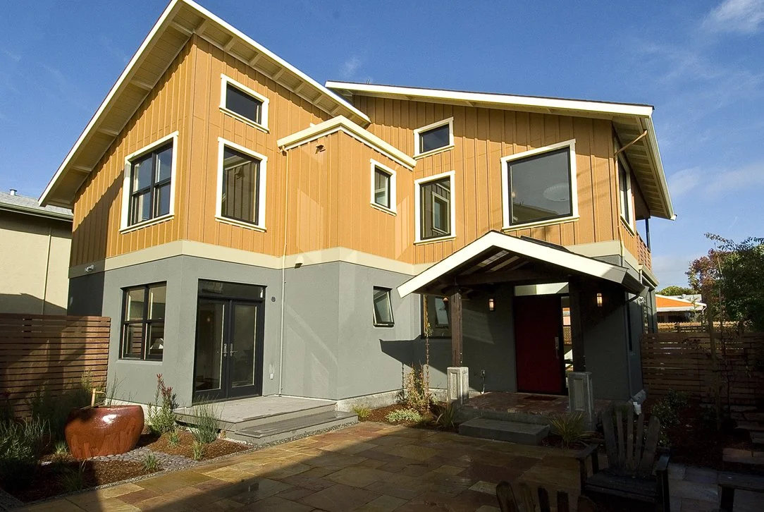 Entry courtyard view showing wood siding, sheltered front door, and hardscape paving that creates a welcoming transition from street to interior living spaces.