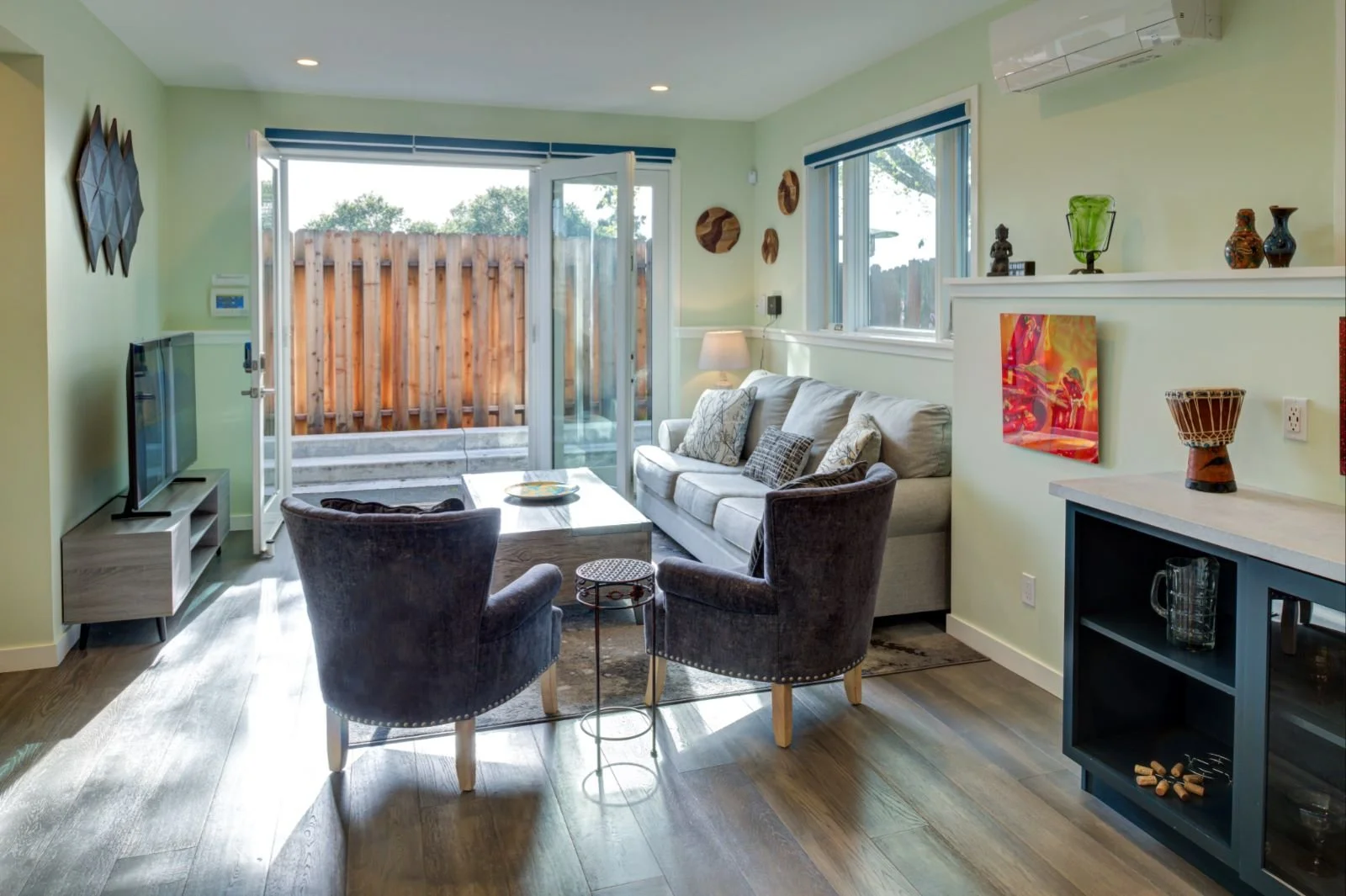 Bright Grand Lake ADU living room by Levitch with sofa, two lounge chairs, wood floors, built in shelving, and glass doors opening to a fenced outdoor patio.