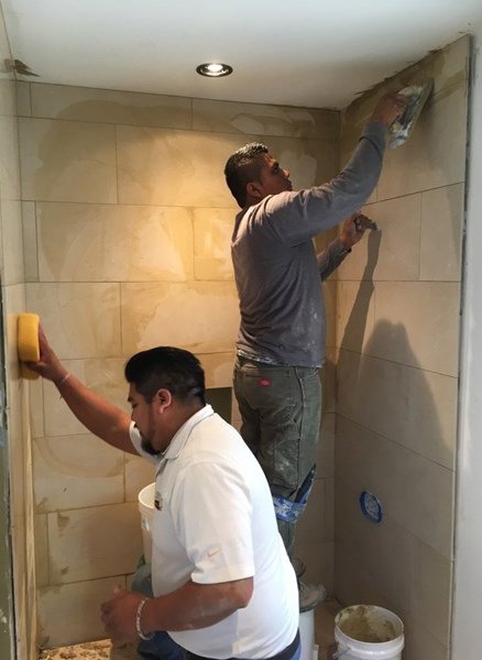 Bathroom remodel in progress with installers setting large format wall tile in the shower and finishing grout lines near the ceiling, part of Levitch Fire and Water updates.