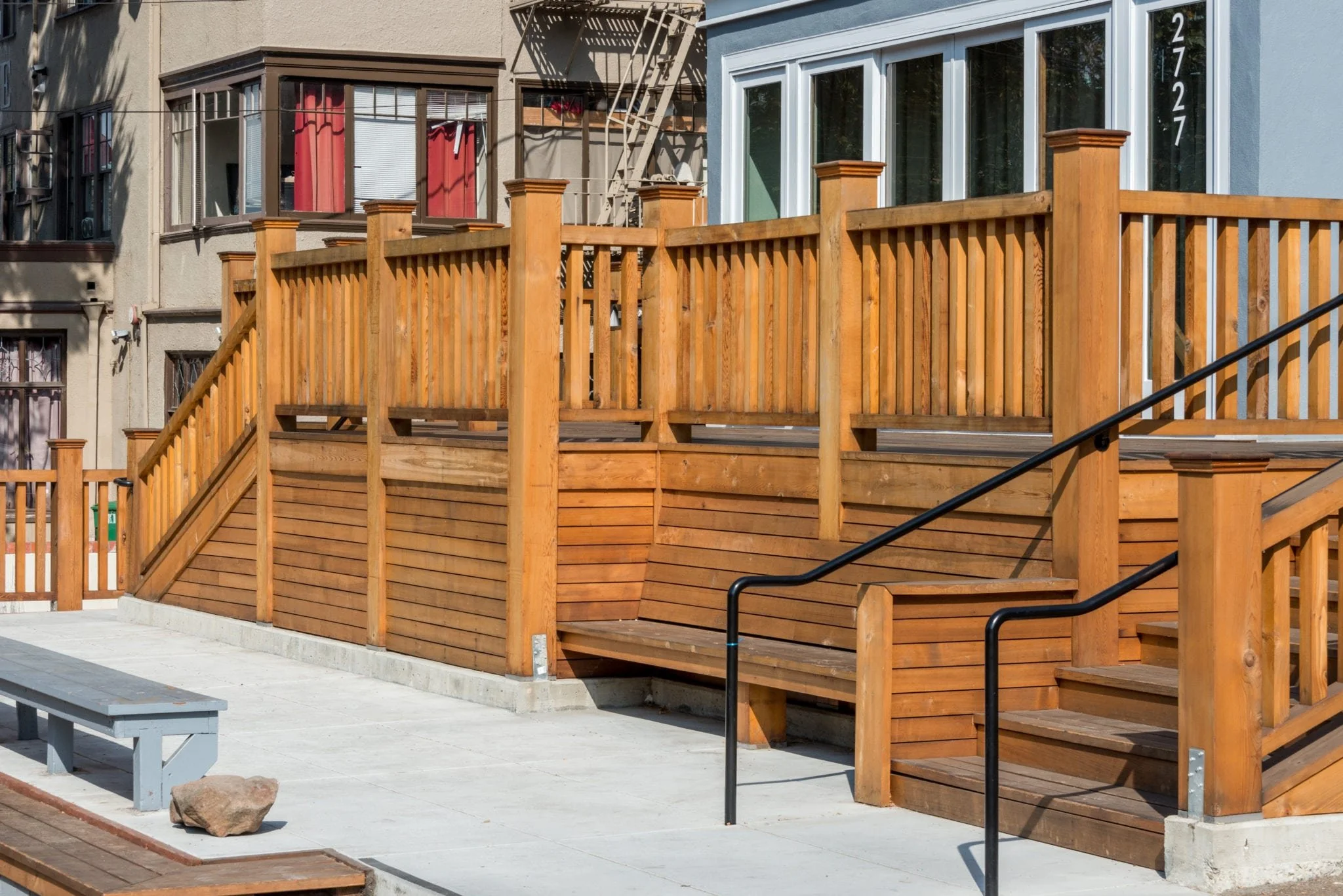 Wood deck with integrated benches and vertical slat railings at an urban fraternity house, creating a welcoming seating edge that connects the building to the sidewalk and street activity.