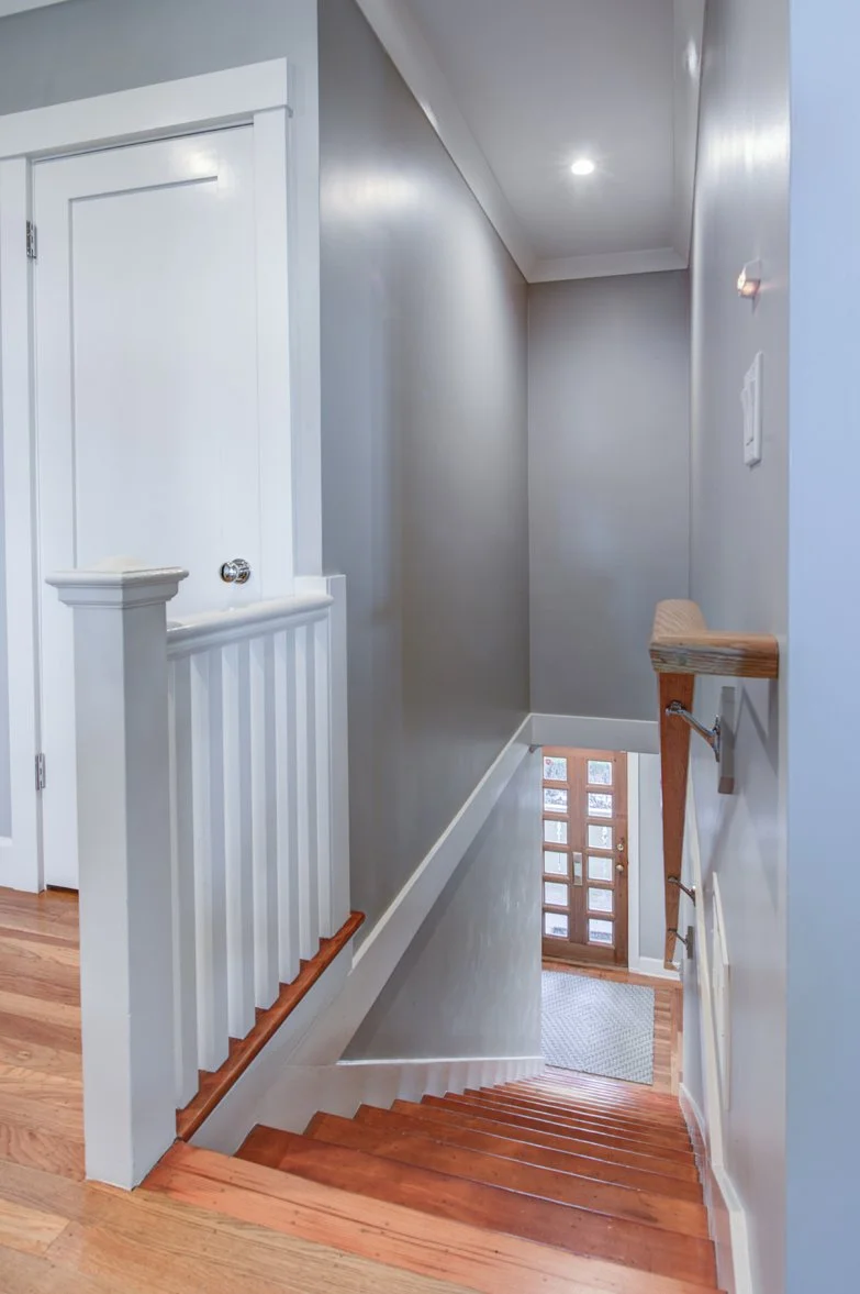 Stair hall with refinished wood steps, white craftsman trim, and a simple wood handrail. Fresh gray walls and bright lighting create a clean transition between floors in this Berkeley home.