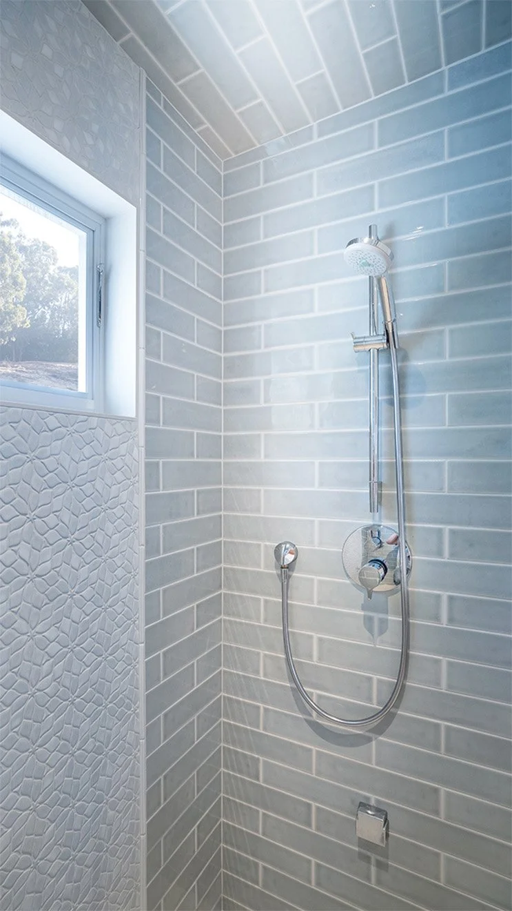 Close-up of a modern tiled shower with soft gray-blue subway tiles and a chrome handheld shower fixture. Natural light filters in through a small window above a section of textured accent tile