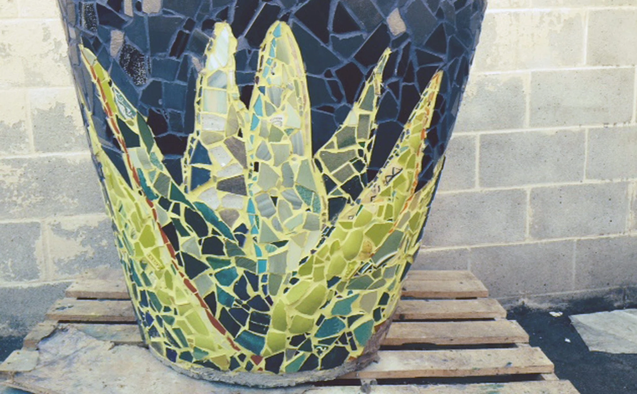 Close-up of mosaic-tiled planter with green and yellow glass pieces on a wooden pallet, detail of parklet artwork
