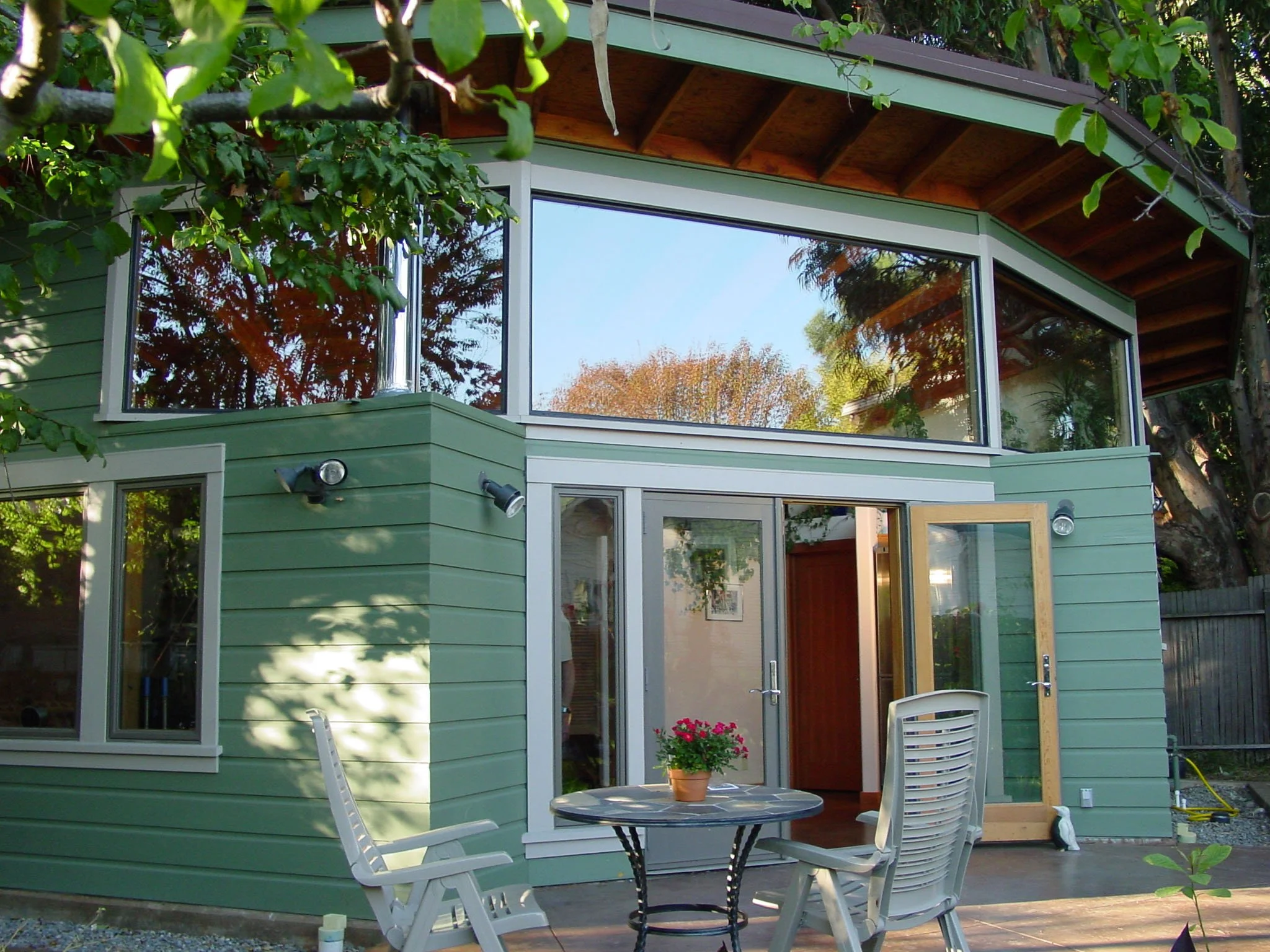 Garden patio view of the one bedroom backyard cottage with outdoor seating, large windows, and open doors, showing indoor and outdoor living areas oriented for passive solar heating.