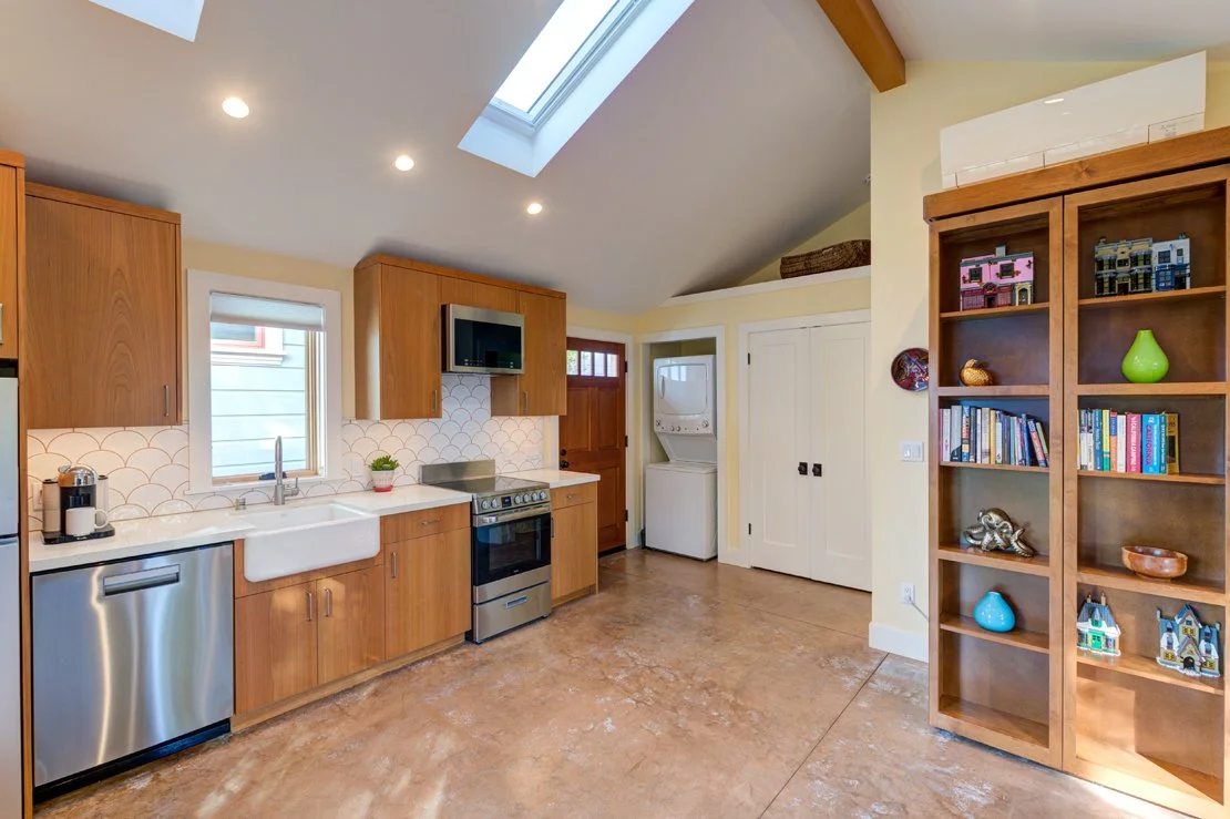 ADU main space with Kitchen wall at the left, vaulted, skylit ceilings, Entry door, stacked Laundry, Bathroom beyond and open living space with Murphy bed.