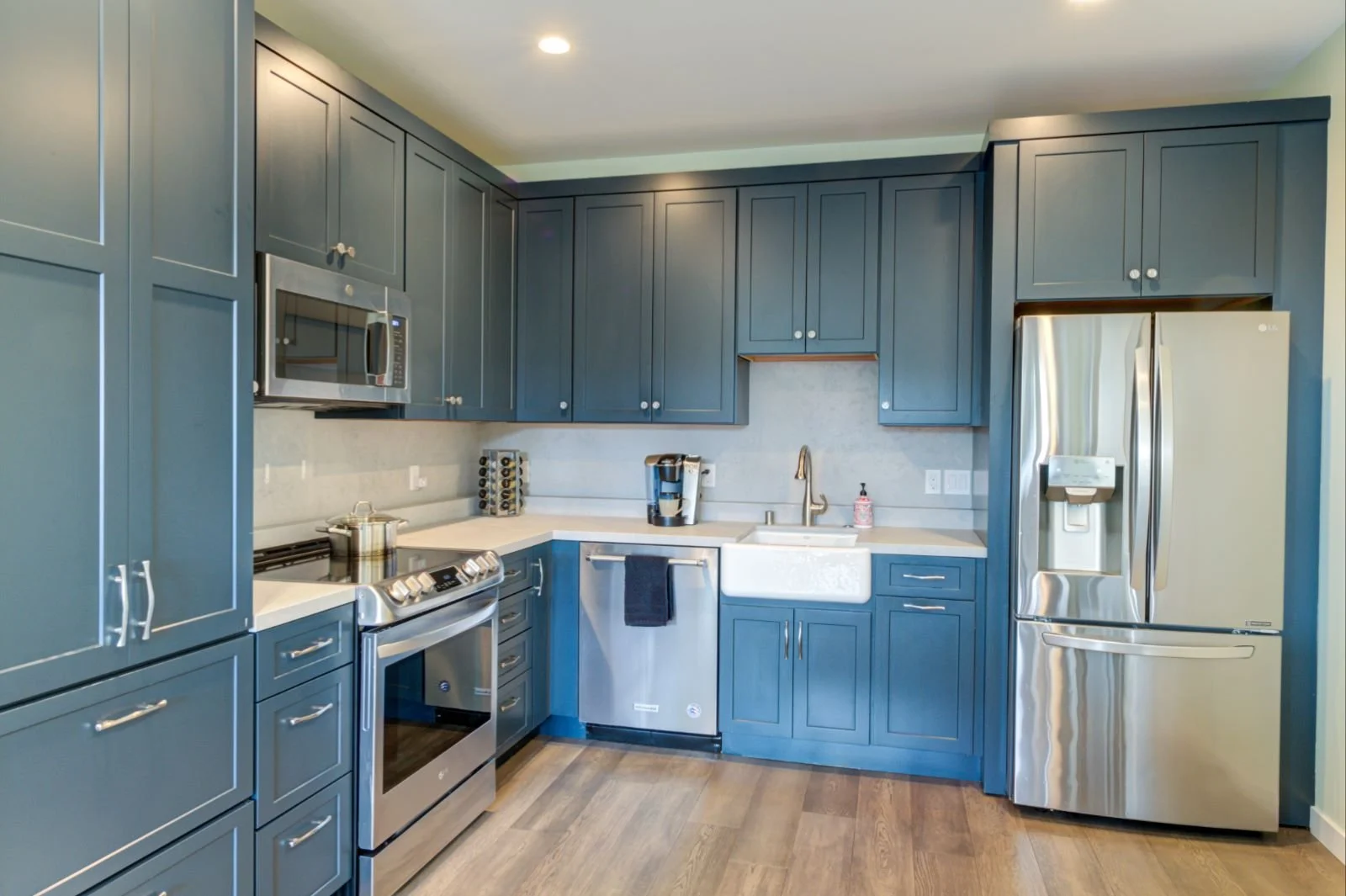 Modern U shaped kitchen in the Grand Lake ADU by Levitch with blue shaker cabinets, stainless appliances, farmhouse sink, refrigerator, and light wood floors.