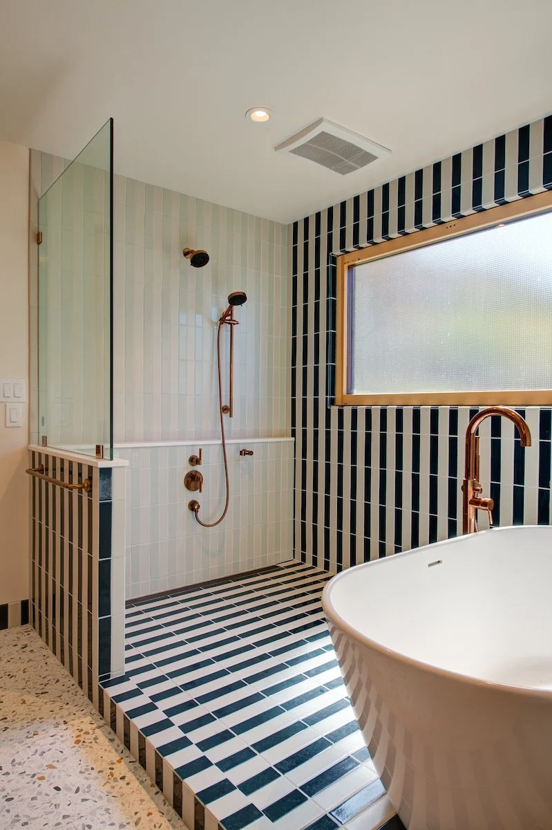 Bold, striped tiles define the shower enclosure. Warm copper fixtures and terrazzo flooring introduce texture and contrast within the bright, contemporary space.
