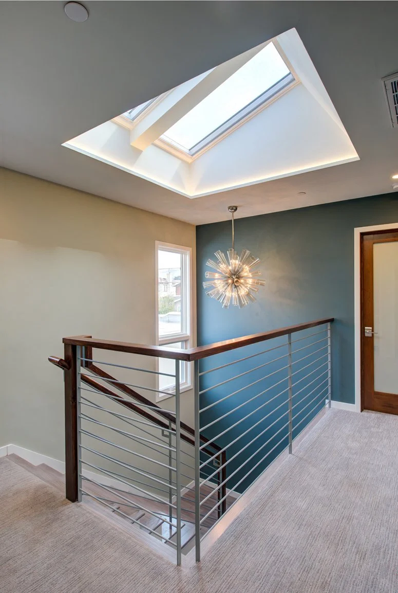 Second-floor stair landing filled with daylight from a large skylight, featuring cable railing, a teal accent wall, and a starburst chandelier above the stair opening.