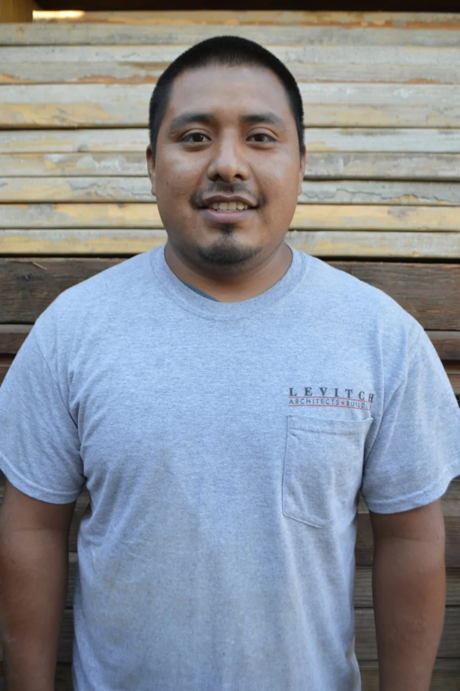 A man with short black hair and a goatee, smiling, standing in front of a wooden fence, wearing a light gray T-shirt with a pocket that has the Levitich Architects logo.
