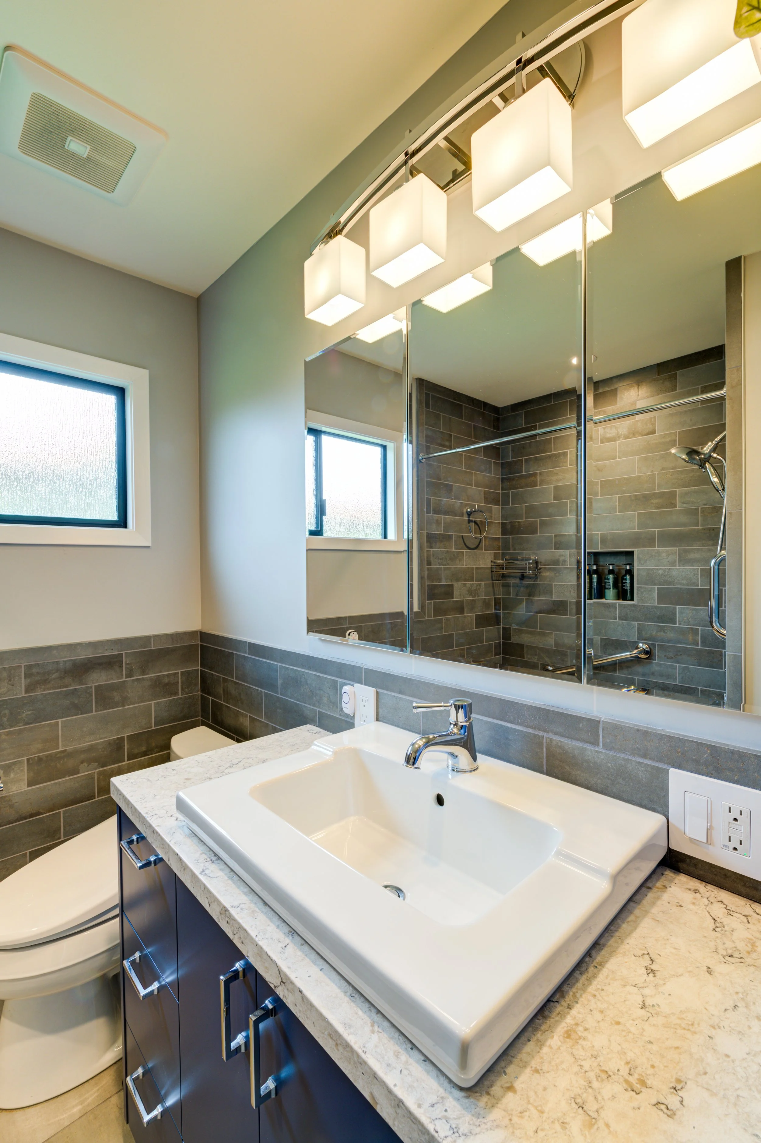 Updated bathroom vanity with blue base cabinets, white countertop and integrated sink, paired with a large mirror, modern lighting, and gray tile walls reflecting a cohesive remodel.