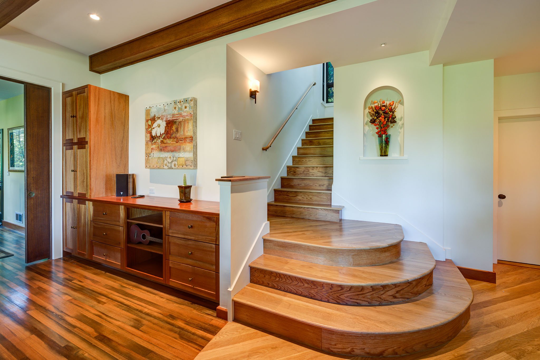 New curved wood stair and built in storage wall leading to the added second story, finished with craftsman trim, warm hardwood floors, and an arched display niche.