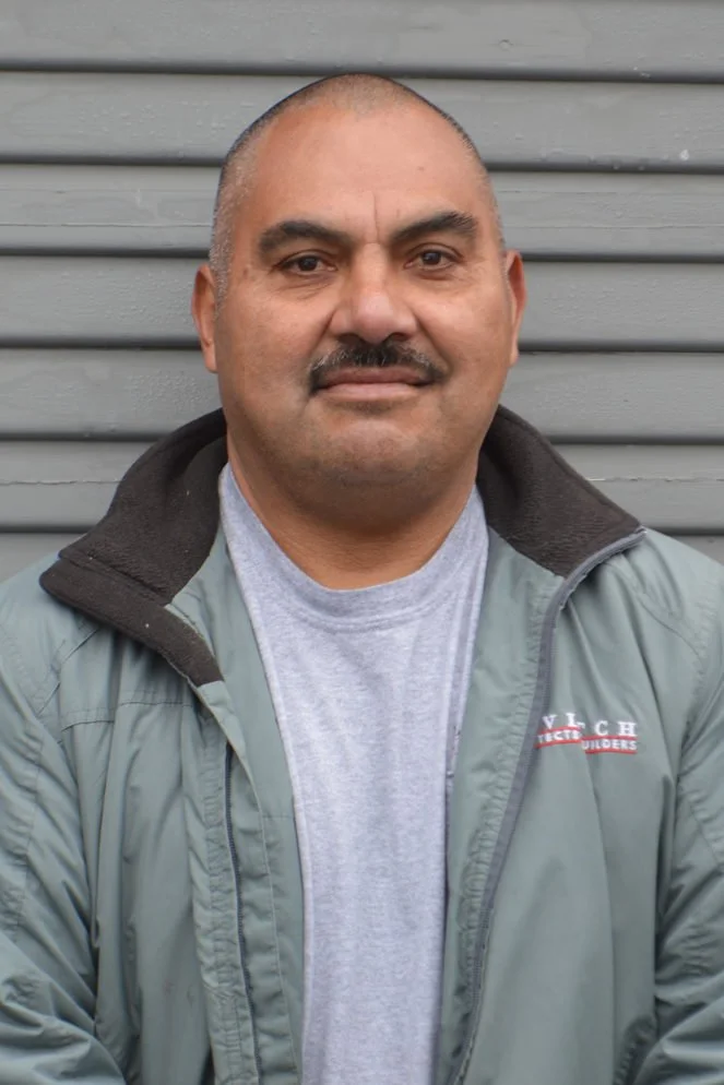 A man with a bald head and a mustache, wearing a gray jacket over a light gray t-shirt, standing in front of a gray, horizontally-paneled wall.