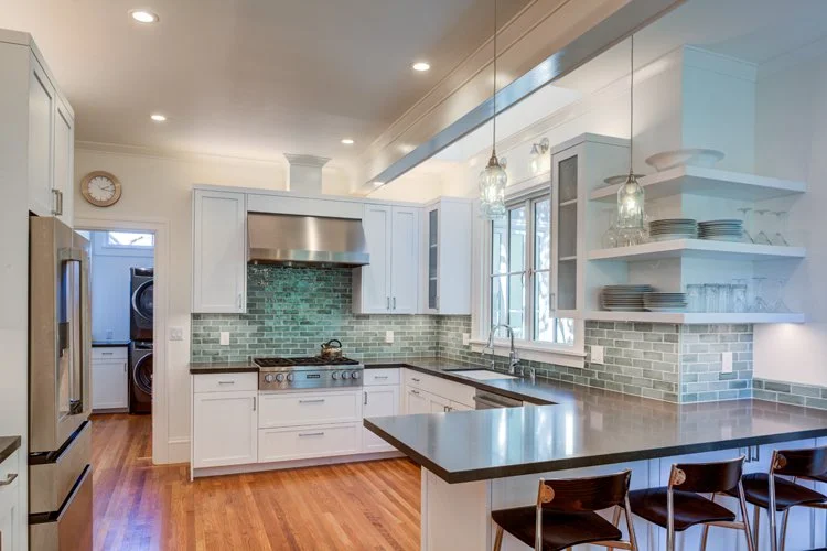 Wide view of the enlarged Julia Morgan kitchen, featuring white cabinetry, a peninsula with seating, a skylit ceiling, and relocated laundry in the background for improved circulation and functionality.