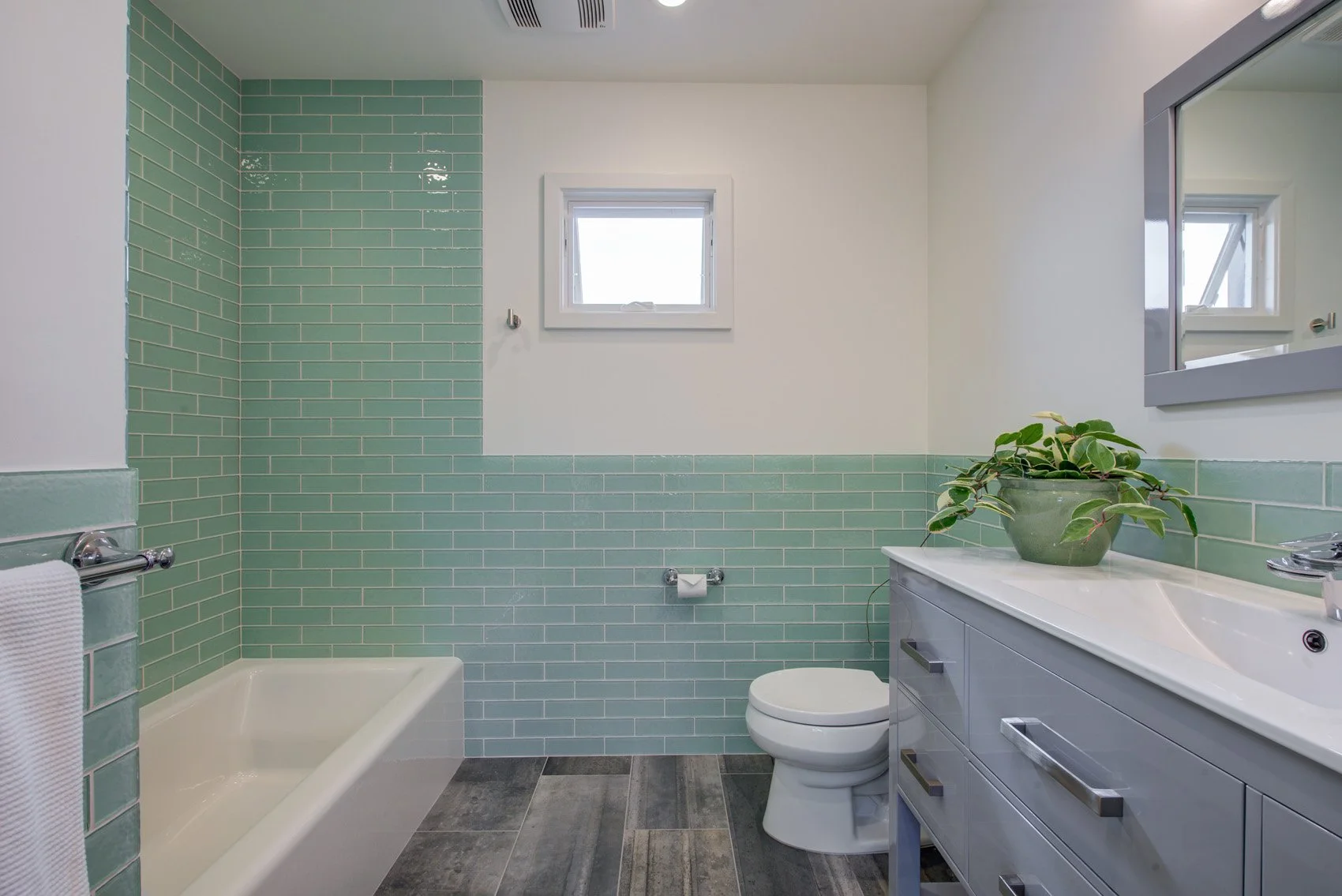 Bathroom with mint green subway tile wainscoting and tub surround, grey vanity with quartz top, small window, and wood look tile flooring that adds warmth to the modern palette.