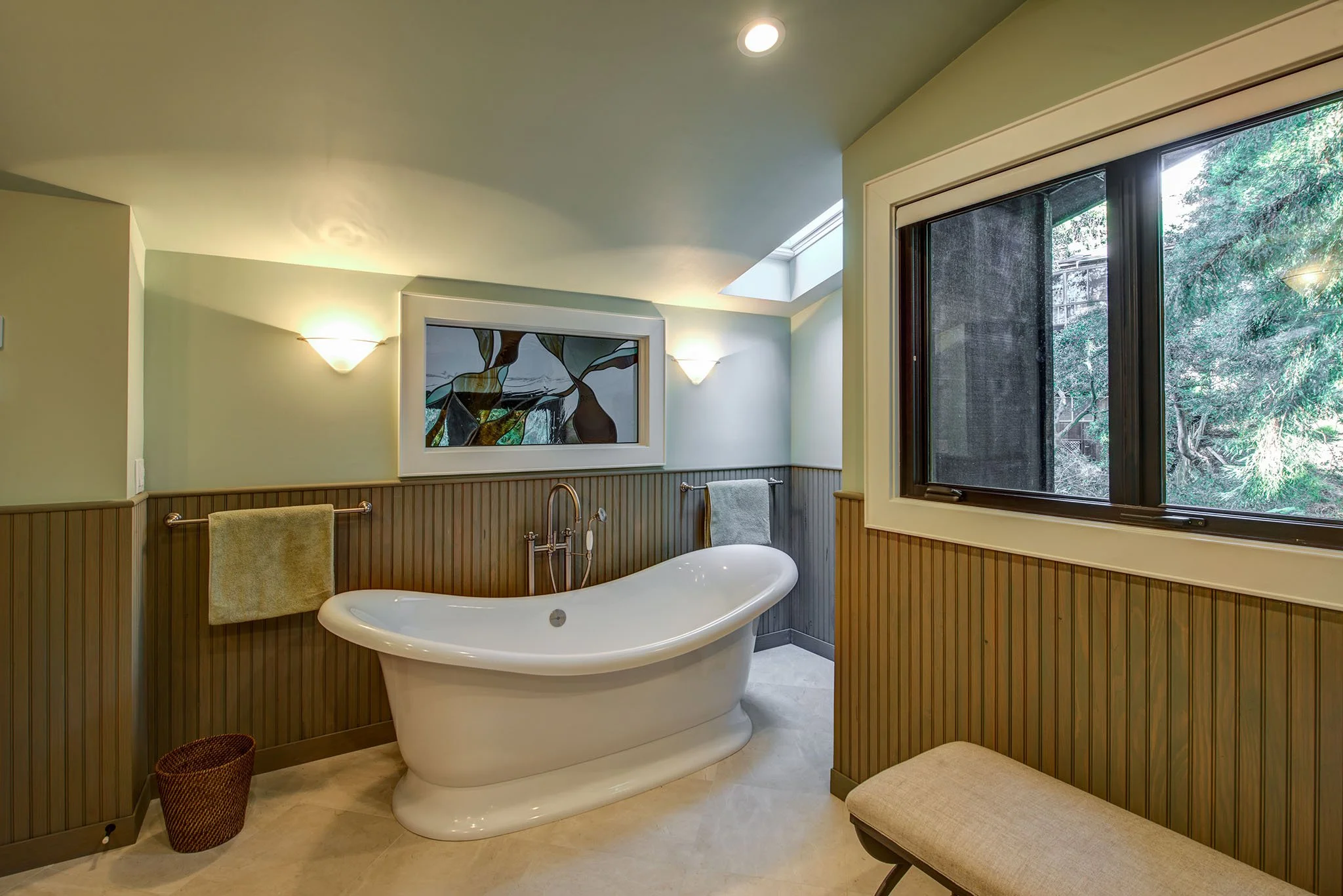 Primary bathroom with a freestanding soaking tub, beadboard wainscoting, and a large window to the trees, offering a calm craftsman retreat after the remodel.