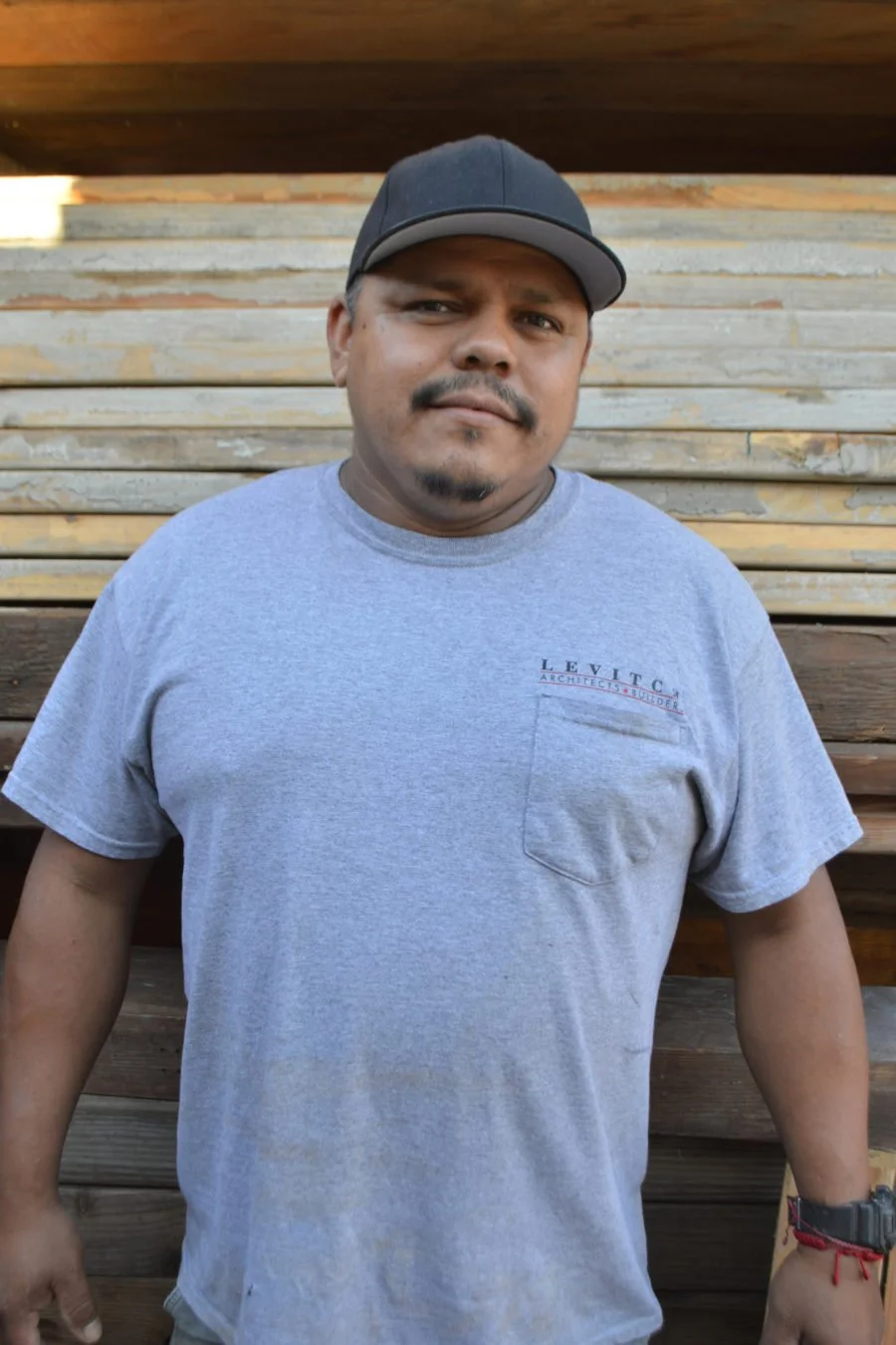 A man wearing a gray t-shirt with a pocket and the words 'LEVTIC ARCHITECTS BUILDER' on it, along with a black baseball cap and a watch, standing in front of a wooden wall.