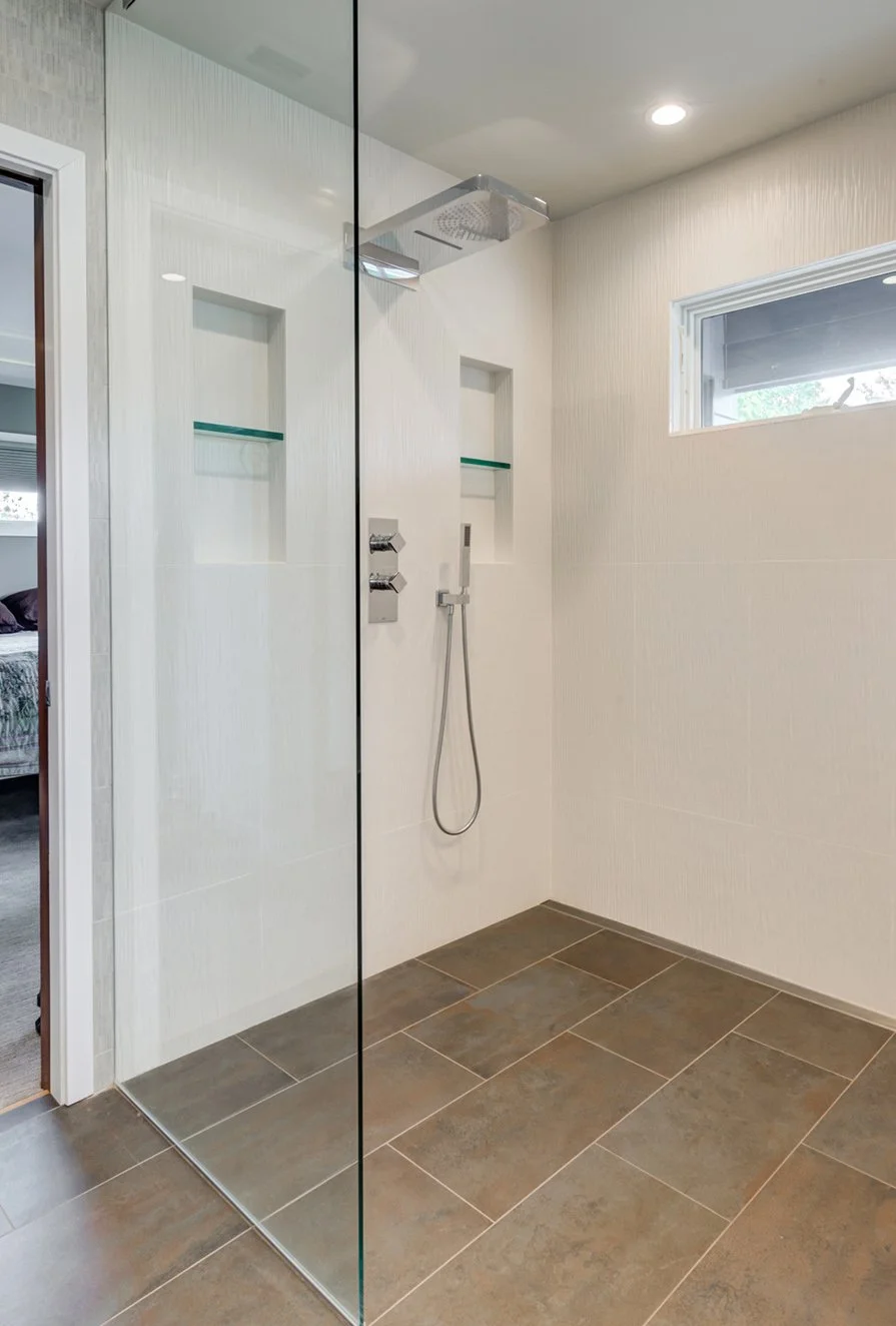 Primary bath walk-in shower with frameless glass, white textured wall tile, built-in niches with glass shelves, rain shower head, and large dark floor tiles for contrast.