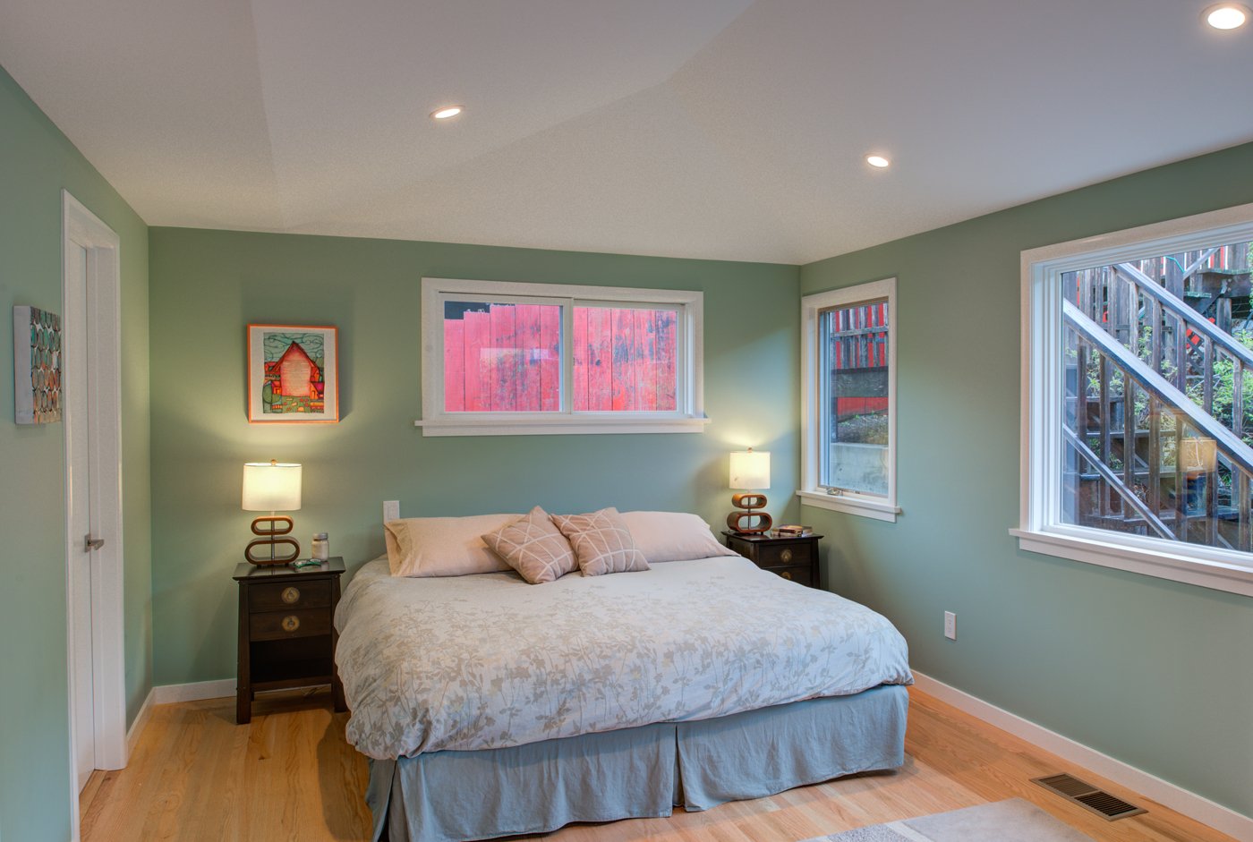 New bedroom suite addition with soft green walls, hardwood floors, and a centered bed between windows, creating a calm primary bedroom retreat in an El Cerrito midcentury home.
