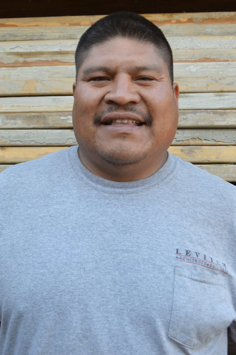A man with short hair and a mustache smiling, wearing a gray t-shirt in front of a wooden wall.