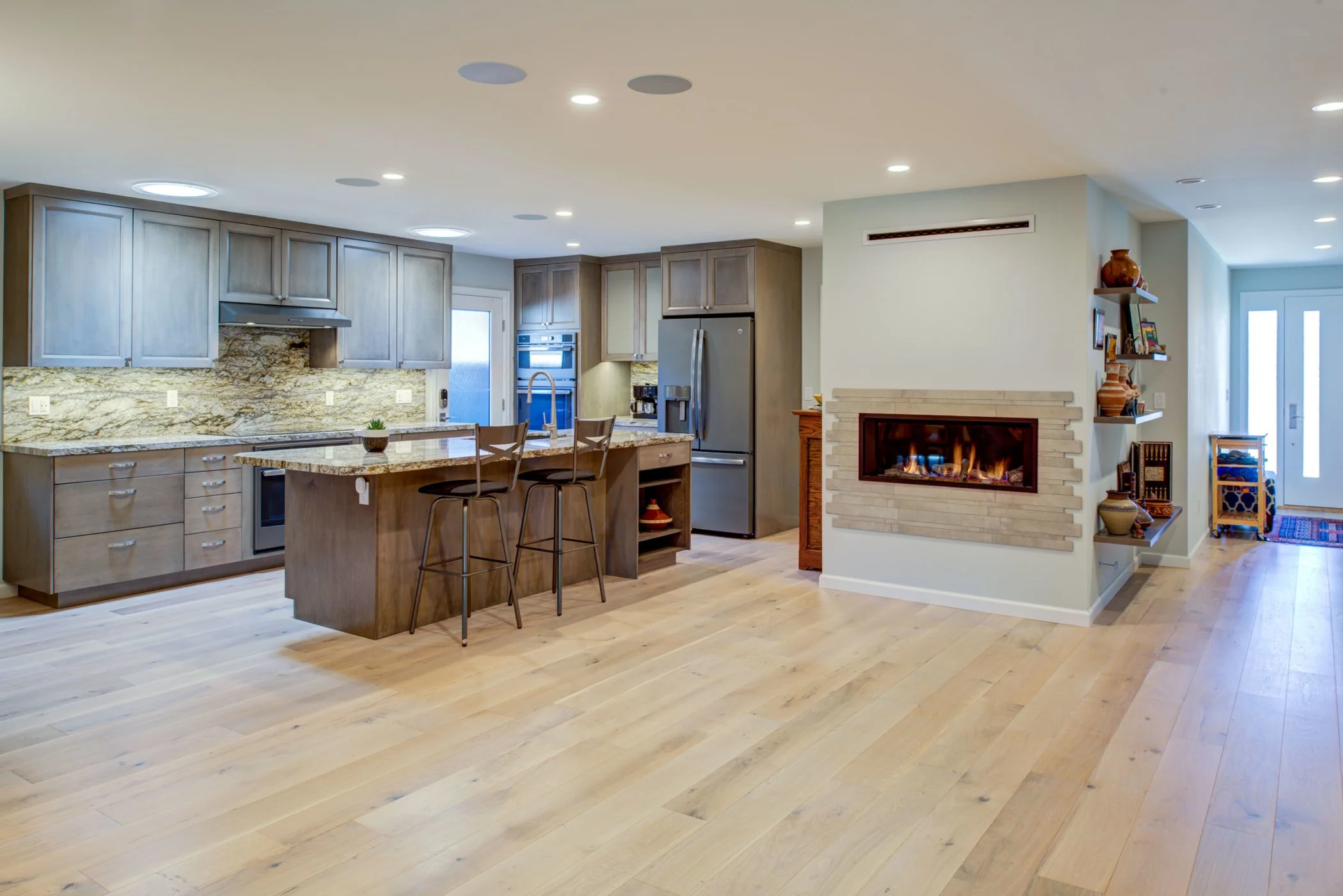 Open living and kitchen area with wide plank wood floors, large island seating, custom cabinetry, and a gas fireplace that connects living and dining spaces.