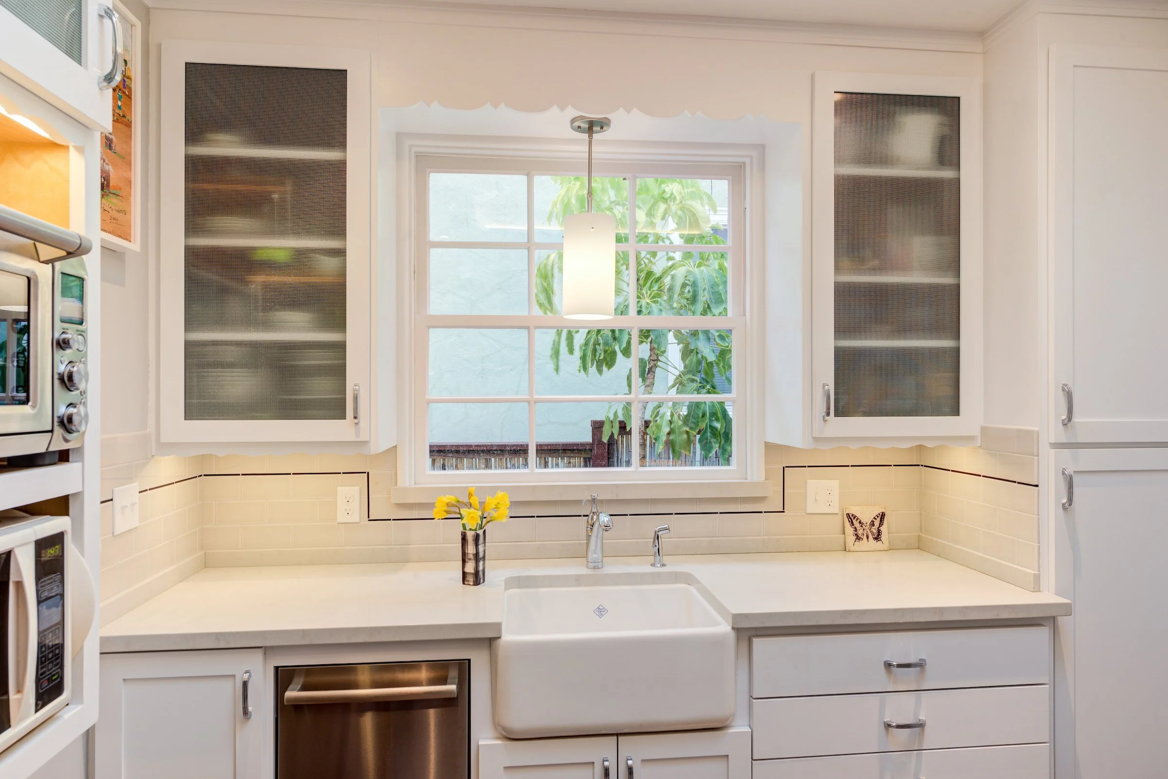 White farmhouse Kitchen sink in white cabinets and two upper cupboards with glass fronts on each side of the window over the sink.