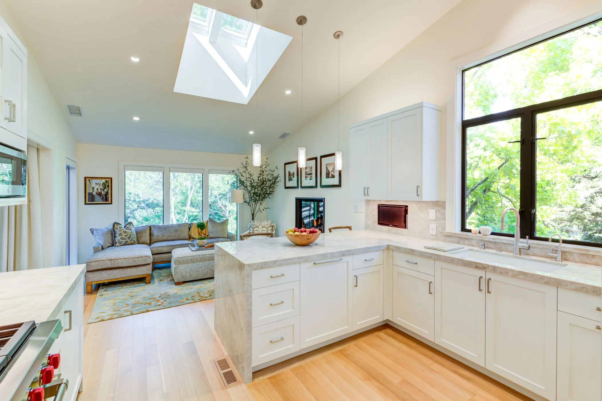 Open kitchen and family room with vaulted ceiling and operable skylights, showing the stone peninsula, pale finishes, and adjacent seating area filled with daylight.