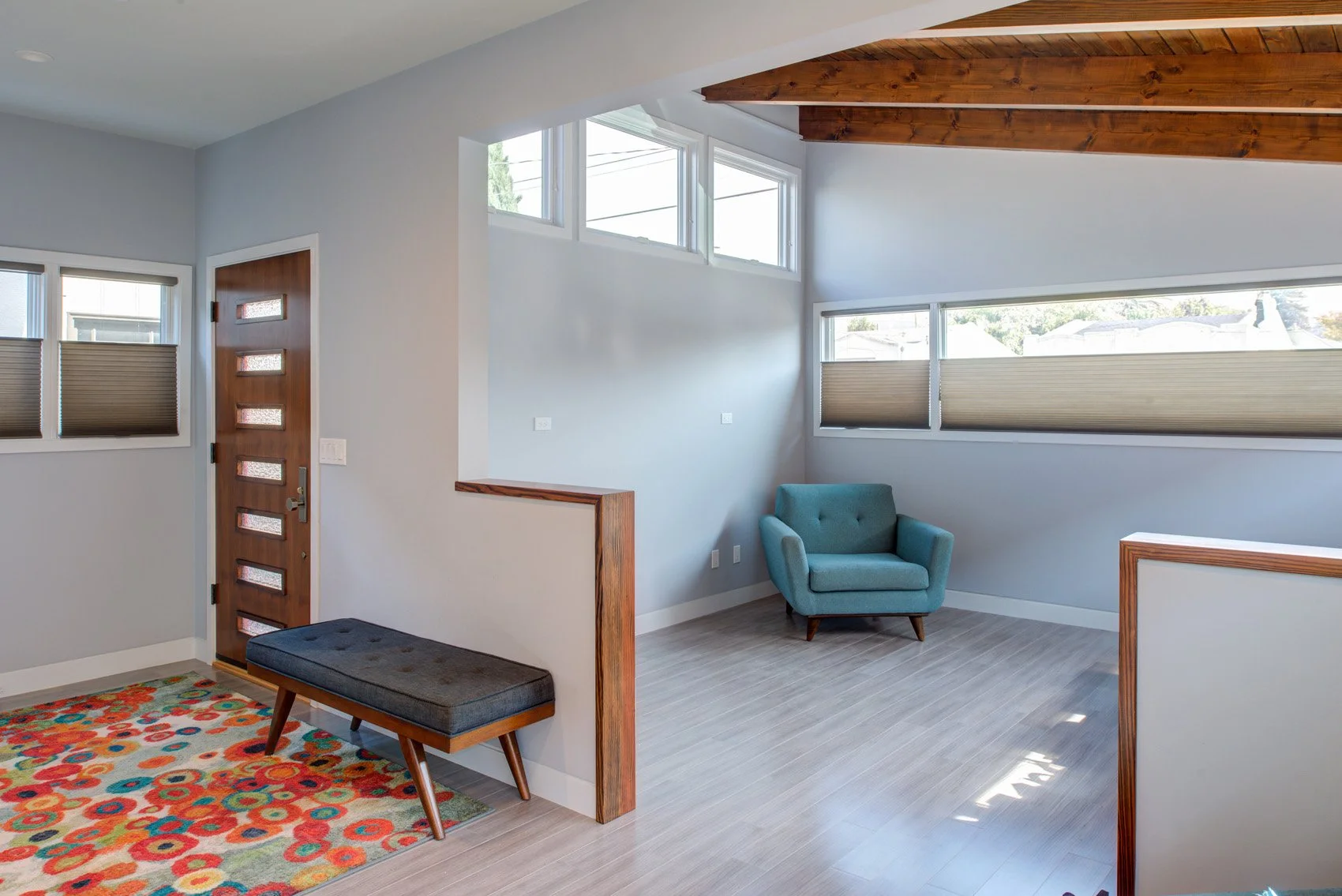 Entry and living area with vaulted ceiling and exposed wood beams, clerestory windows, a warm wood front door, a colorful rug, and a teal lounge chair in soft daylight.