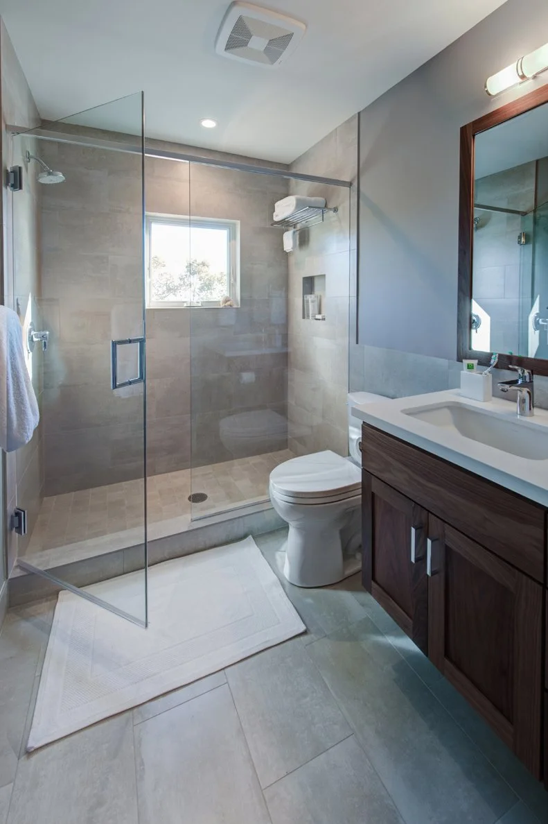 Remodeled bathroom with a frameless glass shower and gray tile walls, plus a floating dark wood vanity and ceiling vent. Cool tones keep the space bright and efficient.