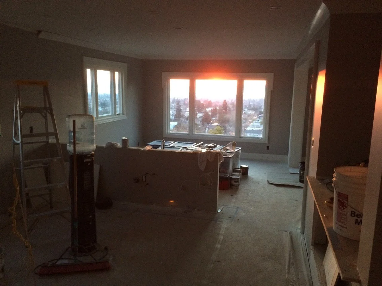 Open plan kitchen and living area mid remodel, with a new island base and tools, looking toward large windows catching sunset light over Berkeley and the Bay.